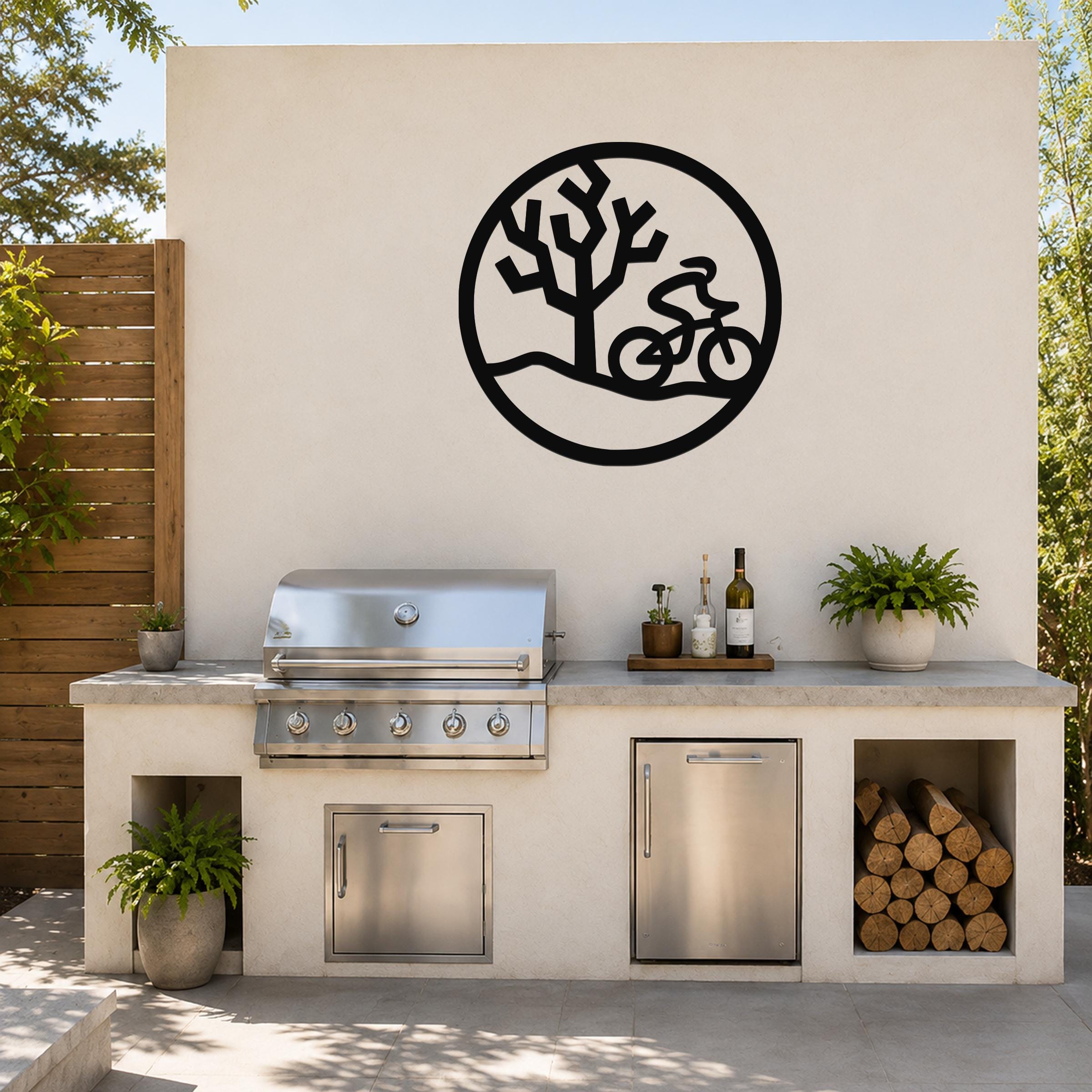 Black metal wall art with a circular silhouette of a cyclist and a Joshua tree, mounted on an outdoor kitchen wall above a stainless steel grill.