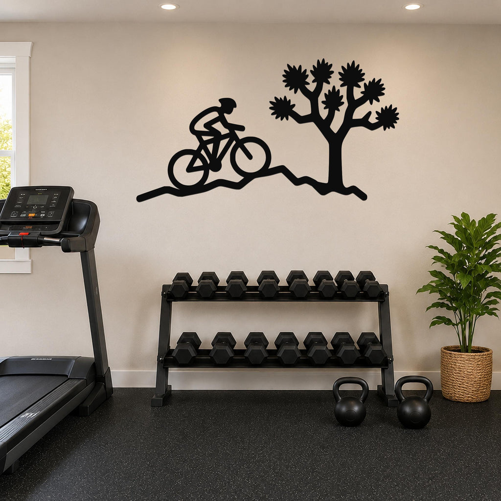 Black metal wall art silhouette of a cyclist on a trail beside a Joshua tree, mounted on a wall above a dumbbell rack in a home gym.