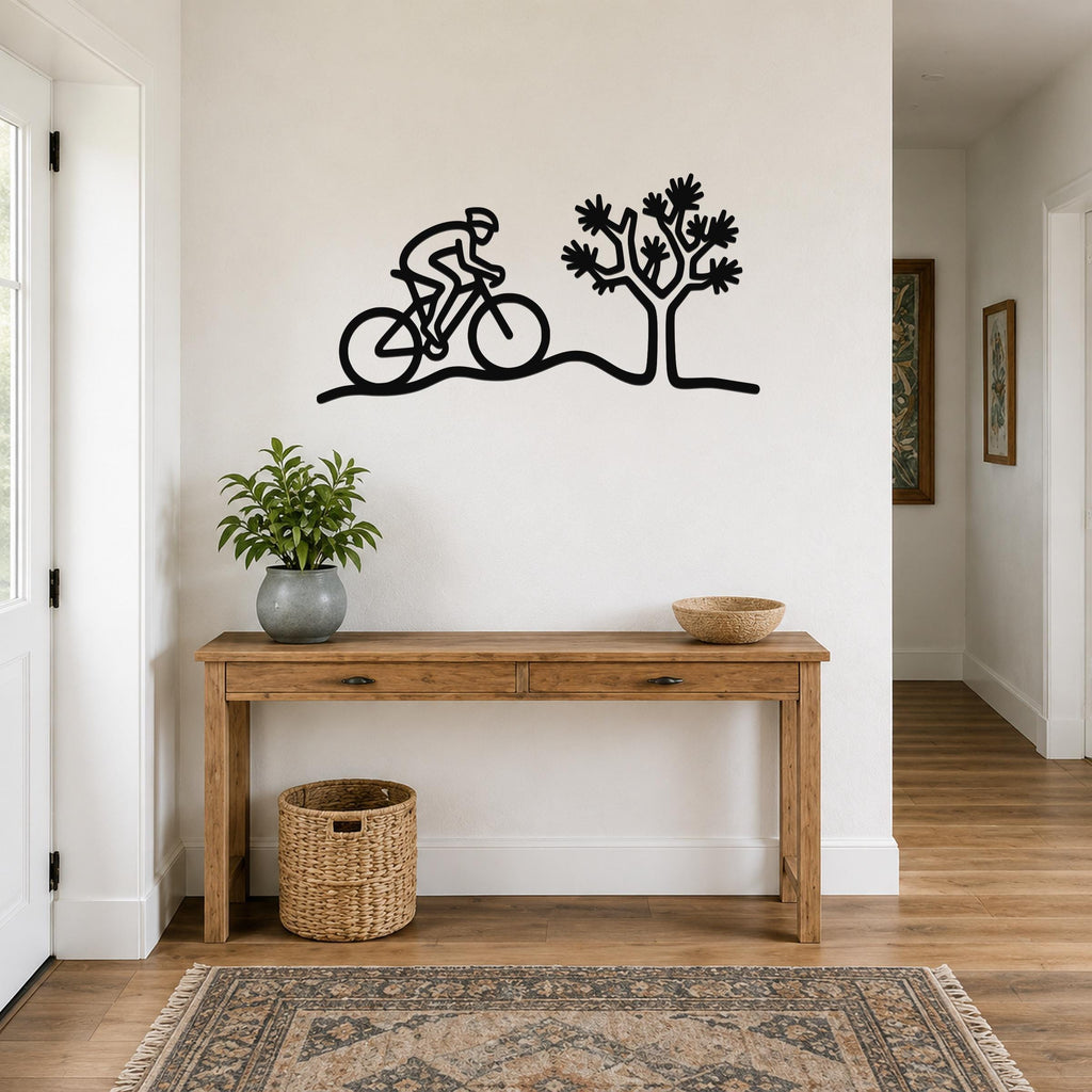 Black metal wall art silhouette of a cyclist riding past a Joshua tree, mounted above a wooden console table in a hallway.
