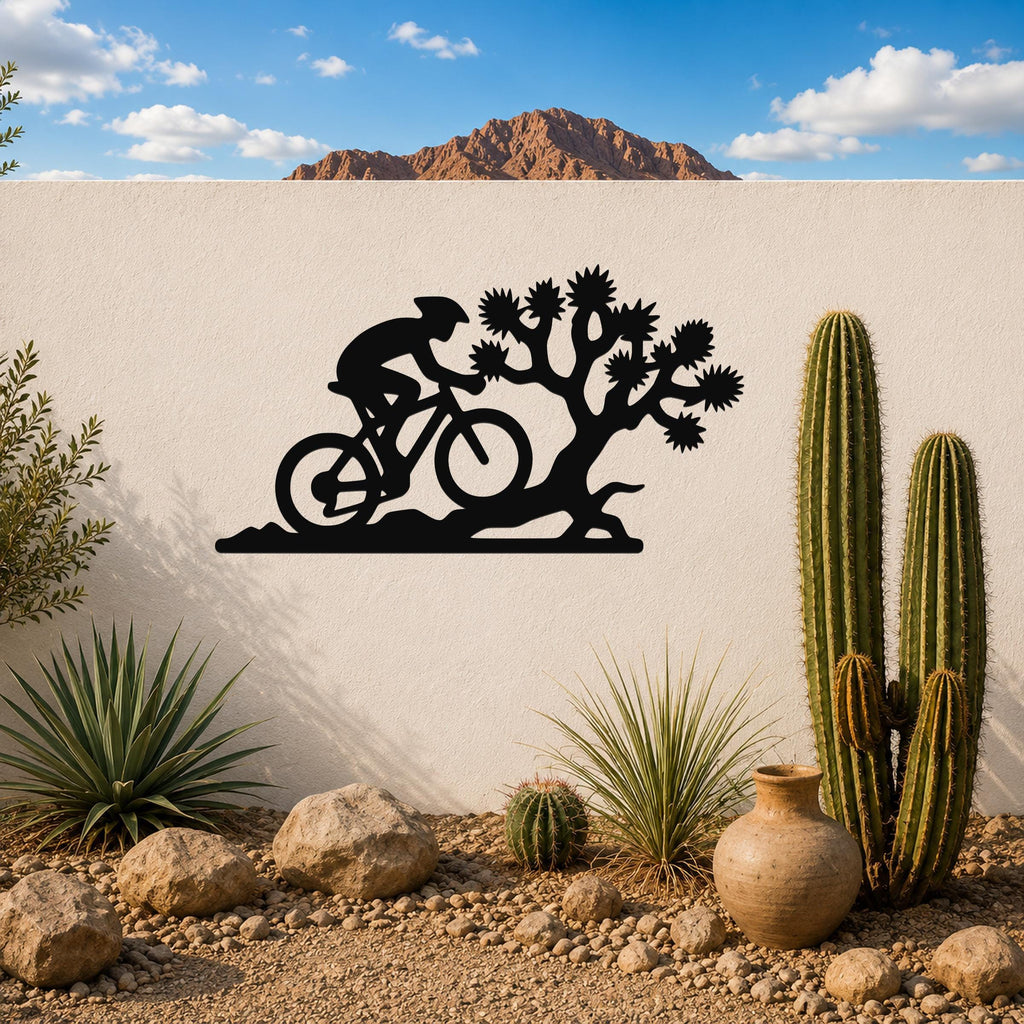 Black metal wall art silhouette of a cyclist and Joshua tree, mounted on an outdoor wall above a desert garden with cacti and rocks.