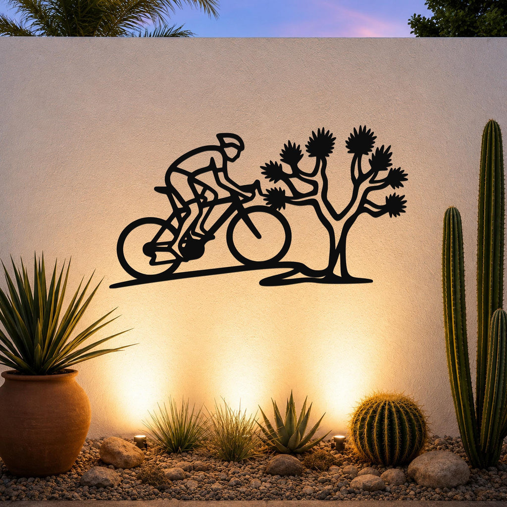 Black metal wall art silhouette of a cyclist and Joshua tree, mounted on an outdoor wall above a desert garden with cacti and warm uplighting.