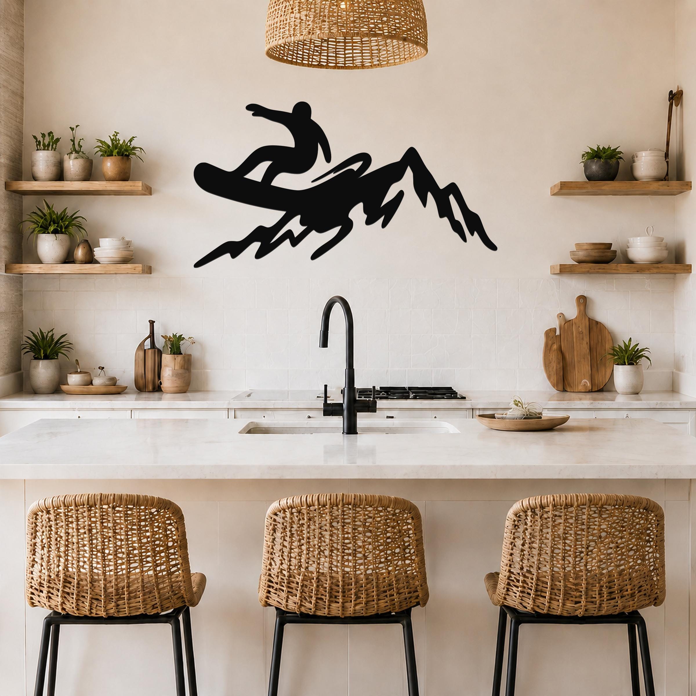 Black metal wall art silhouette of a snowboarder on a mountain, mounted above a kitchen counter with wicker bar stools and open shelves.