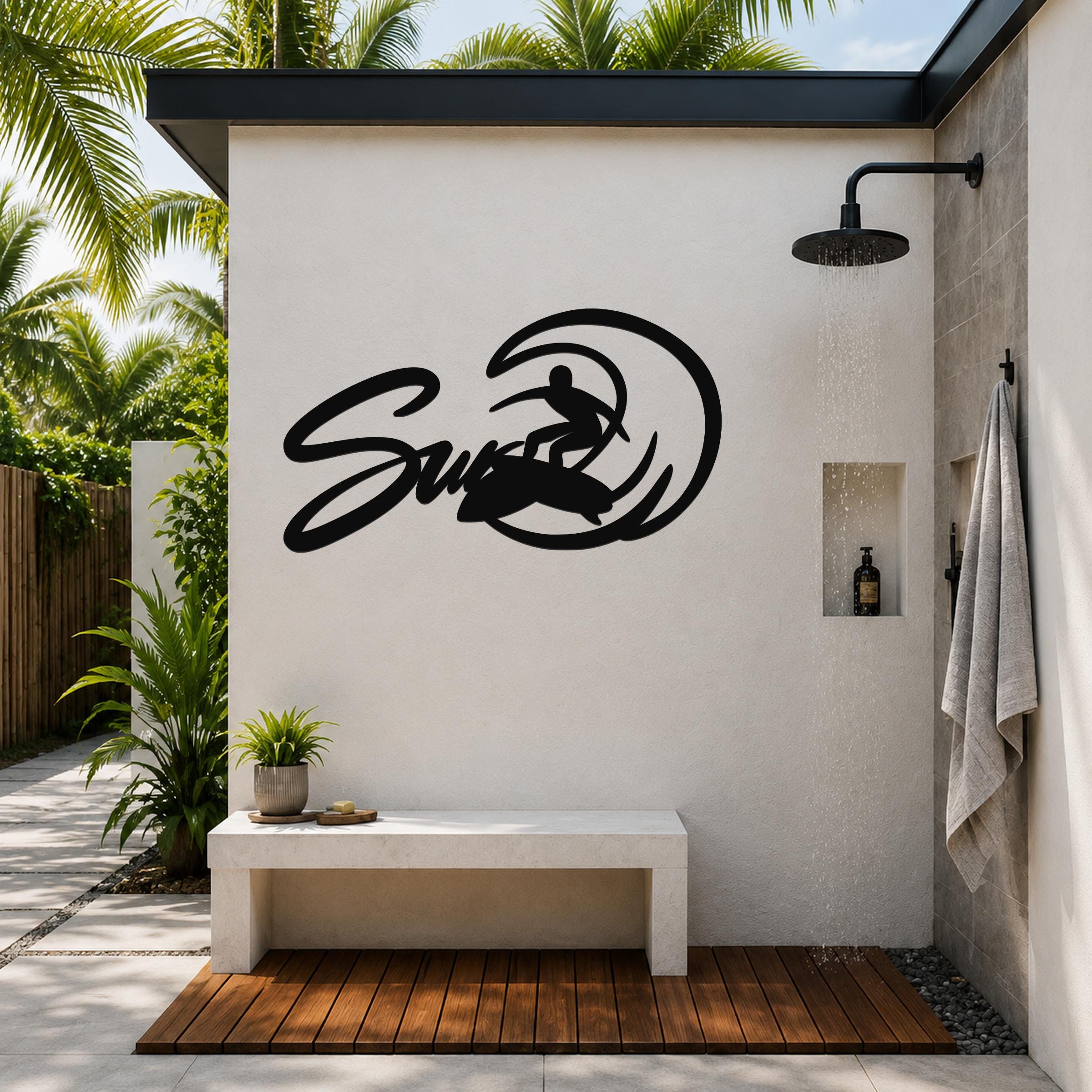Black metal wall art featuring a surfer silhouette riding a wave, mounted on an outdoor shower wall above a concrete bench.