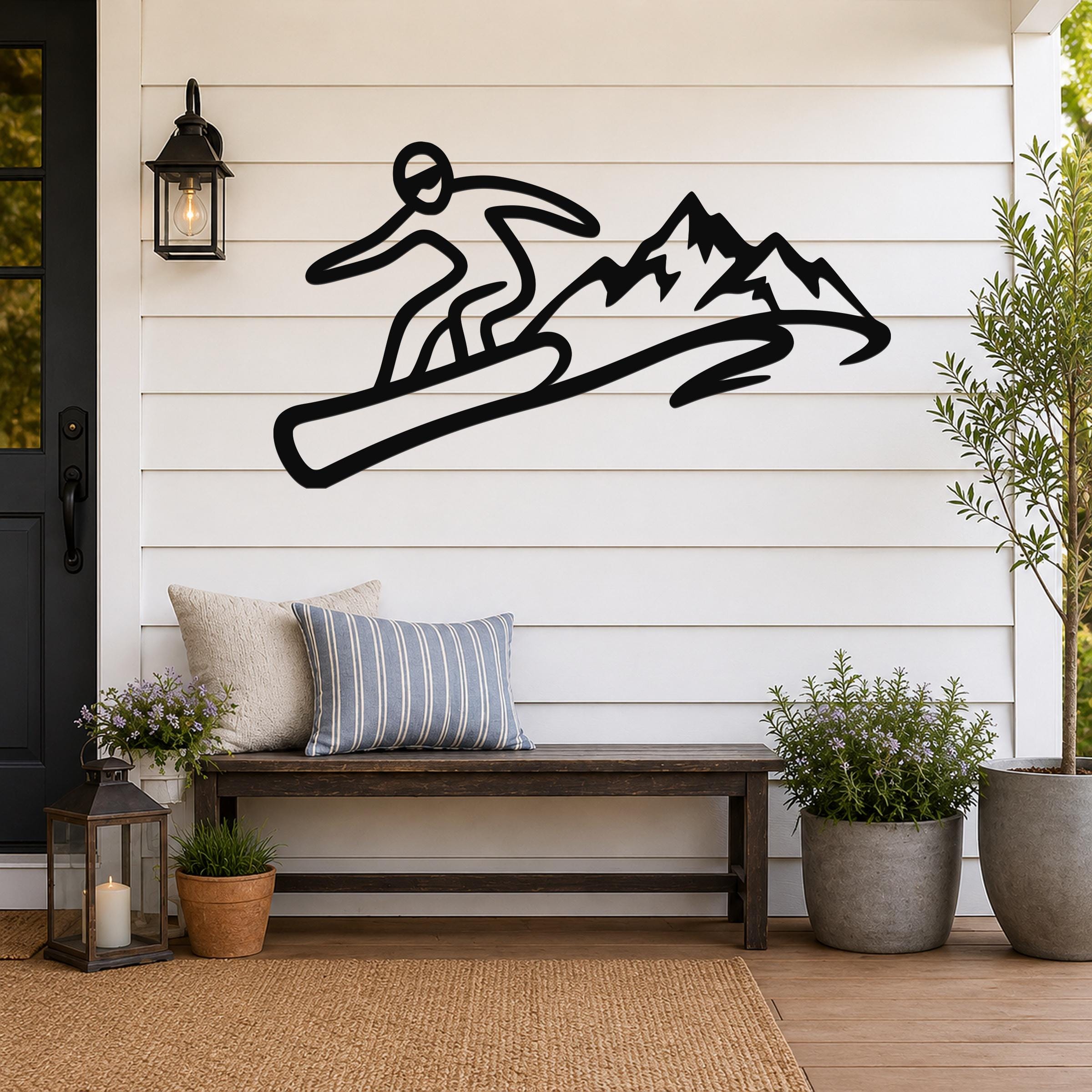 Black metal wall art featuring a snowboarder silhouette in motion above a mountain range, mounted on a white shiplap wall above a wooden bench on a porch.