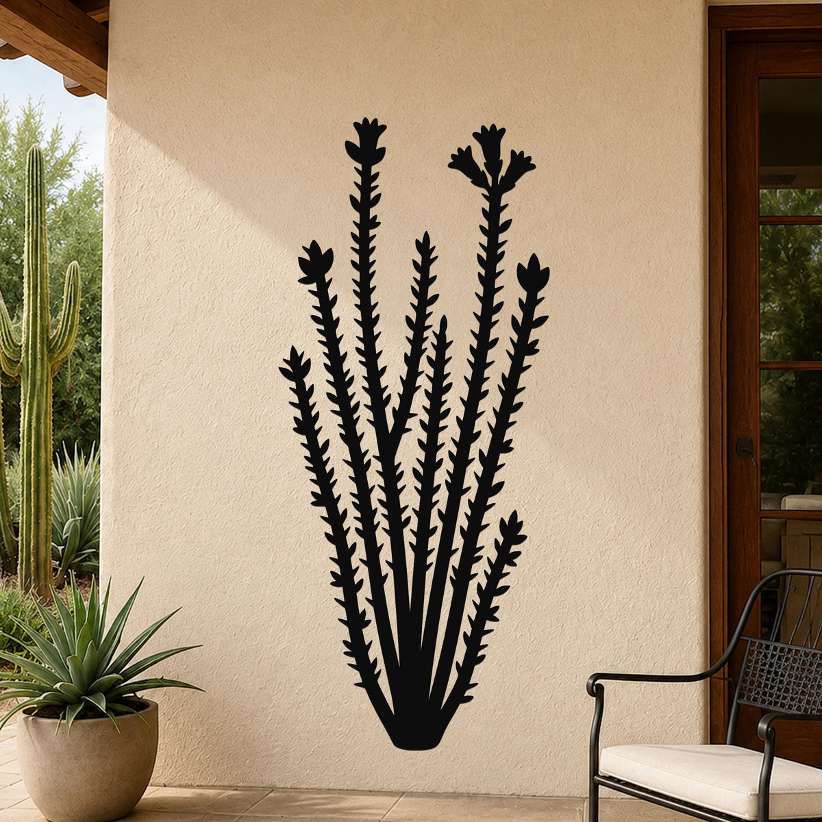 Black metal ocotillo wall art with spiny stems and flower cutouts, mounted on a beige outdoor wall beside a patio chair and potted plant.