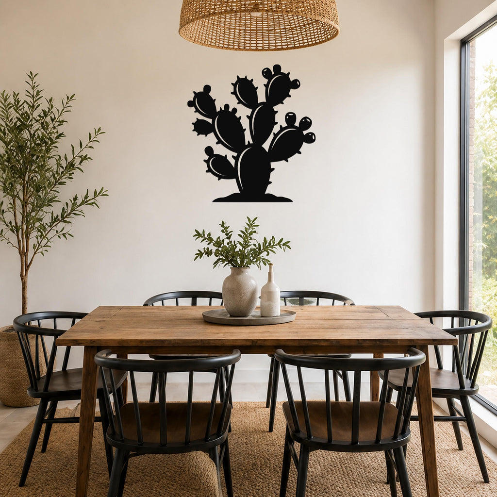 Black metal cactus wall art with a silhouette of a prickly pear, mounted on a white wall above a wooden dining table in a bright room.