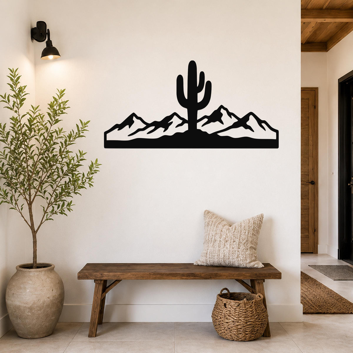 Black metal wall art featuring a saguaro cactus and mountain silhouette, mounted on a white wall above a wooden bench in a hallway.