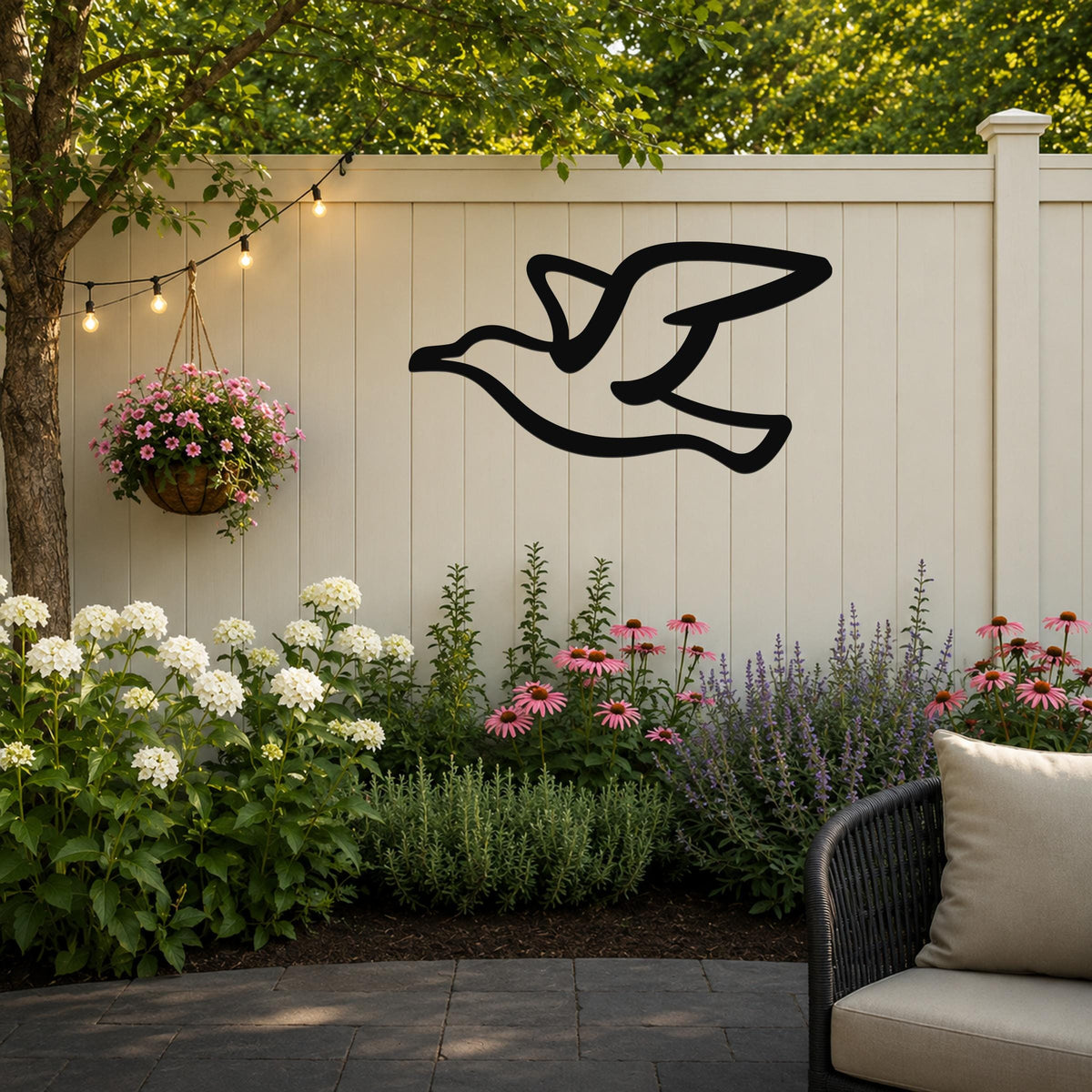 Black metal seagull wall art silhouette mounted on a light-colored outdoor fence, above a garden bed with flowers and string lights.