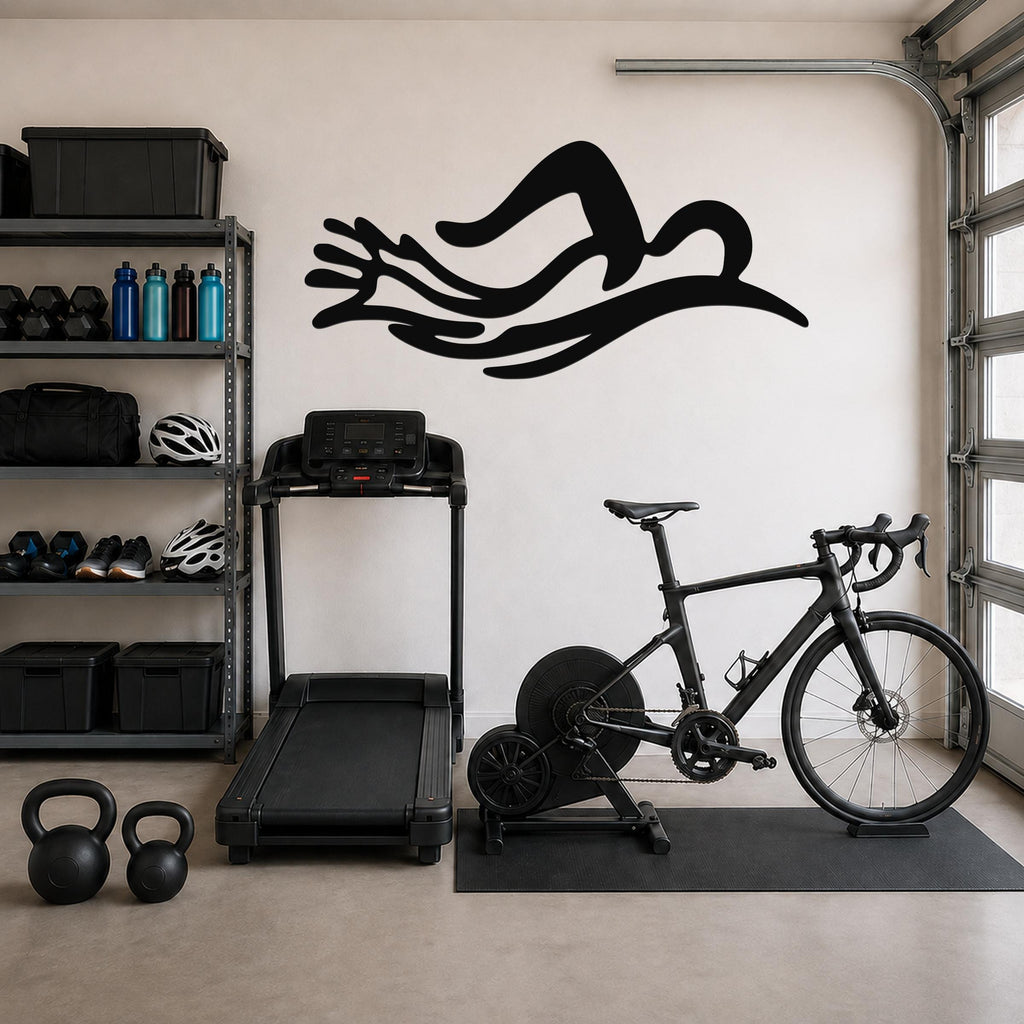 Black metal wall art in the shape of a swimmer's silhouette with flowing motion lines, mounted on a garage wall above a treadmill and exercise bike.