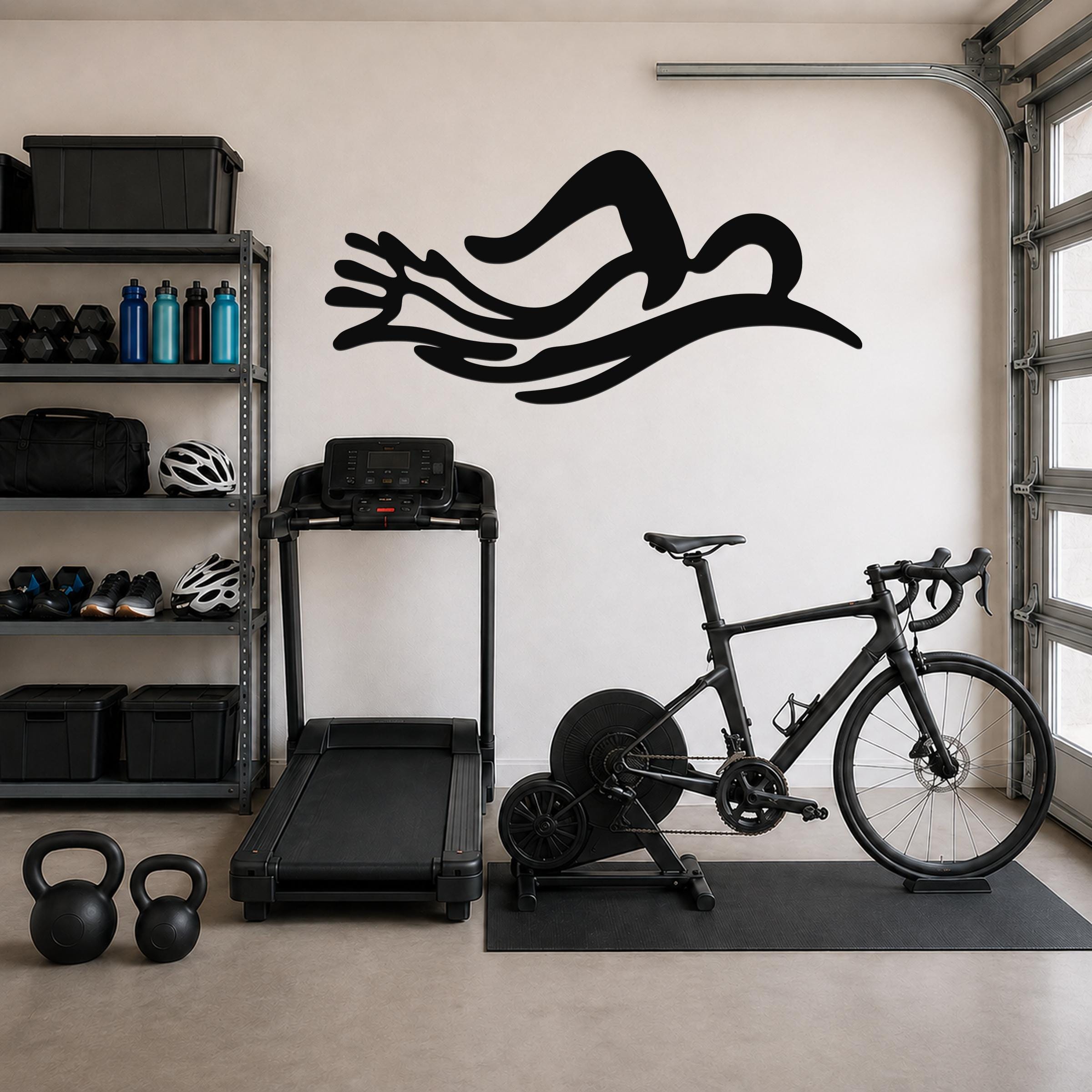 Black metal wall art in the shape of a swimmer's silhouette with flowing motion lines, mounted on a garage wall above a treadmill and exercise bike.