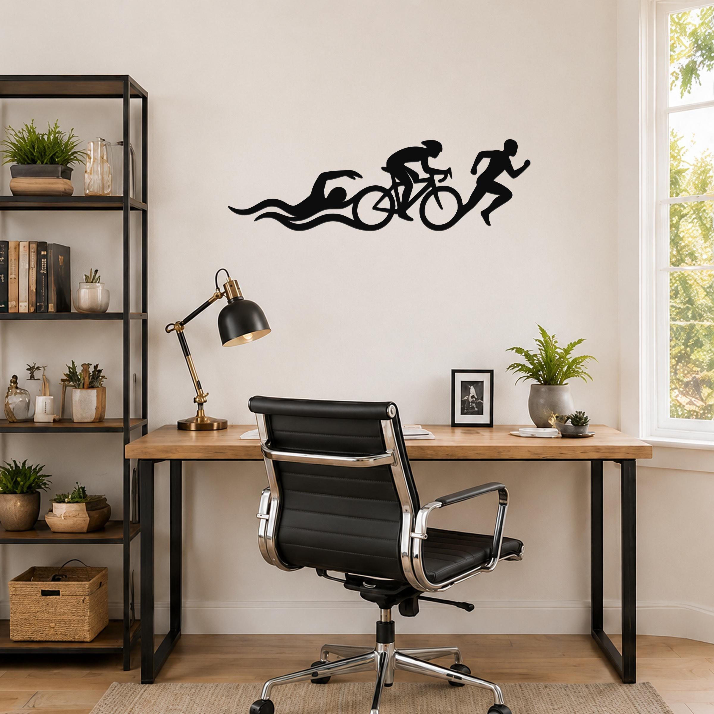 Black metal triathlon wall art with swim, bike, and run silhouettes and motion lines, mounted above a wooden desk in a home office.