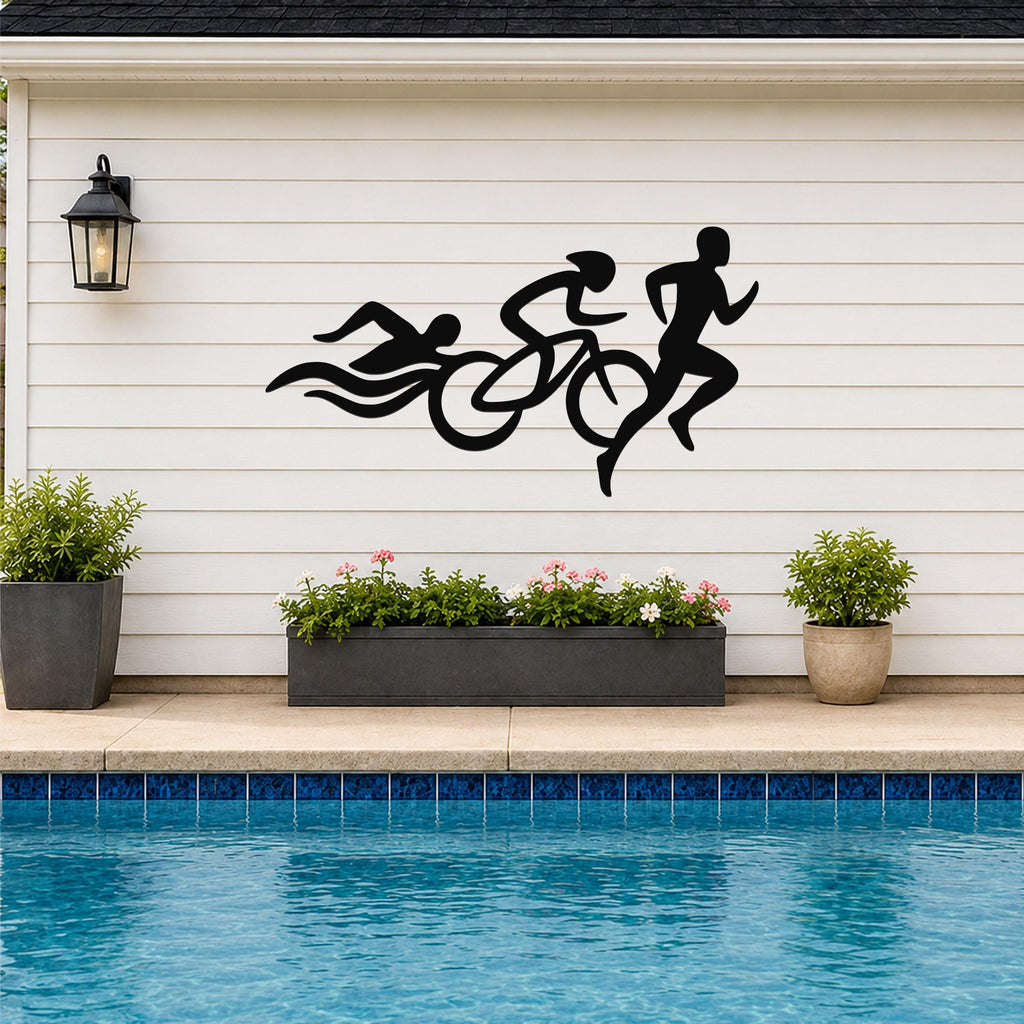 Black metal triathlon wall art with swim, bike, and run silhouettes, mounted on a white siding wall above a poolside planter.