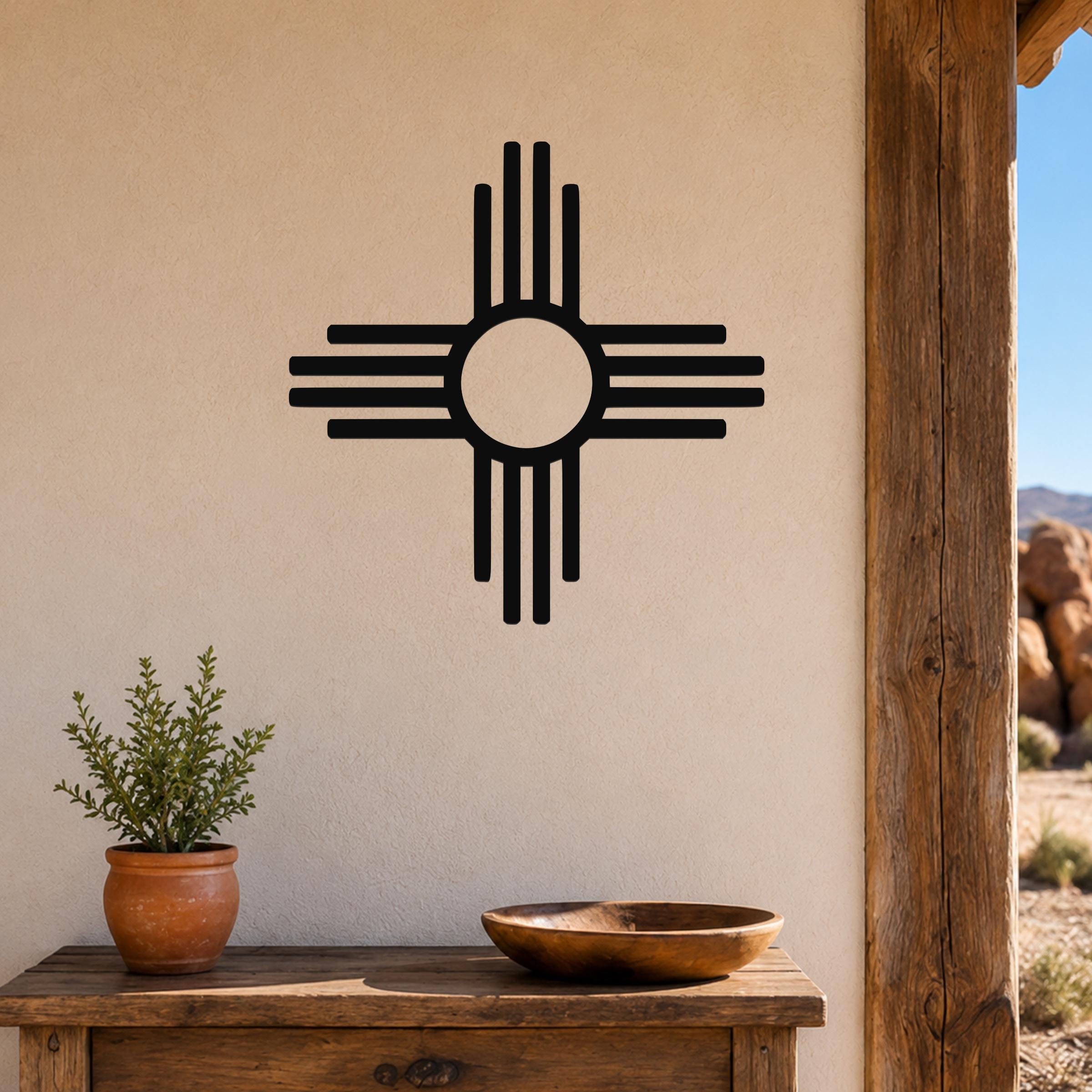 Black metal Zia Sun symbol wall art with a bold circular design and radiating lines, mounted on a beige stucco wall above a wooden table with a potted plant and bowl.