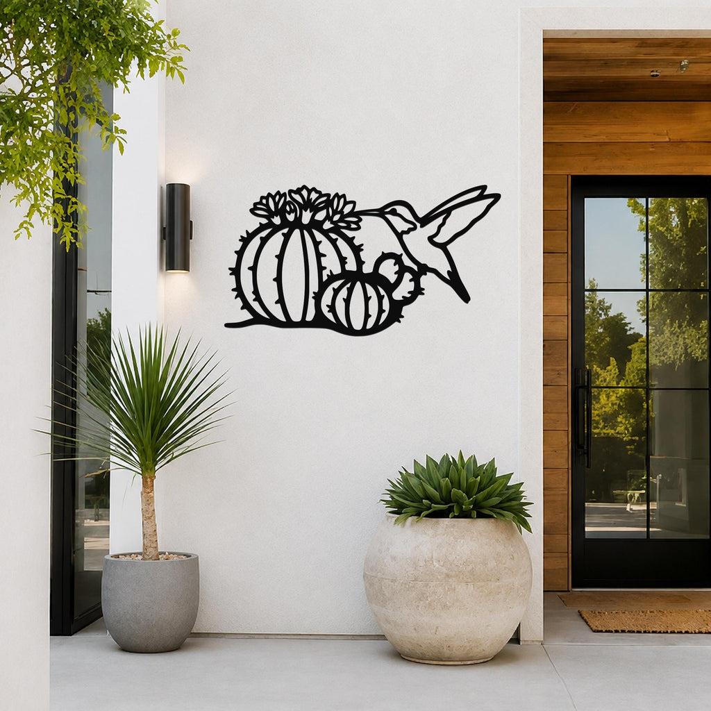 Black metal wall art featuring a hummingbird and barrel cactus with flower cutouts, mounted on a white exterior wall near a glass door and potted plants.