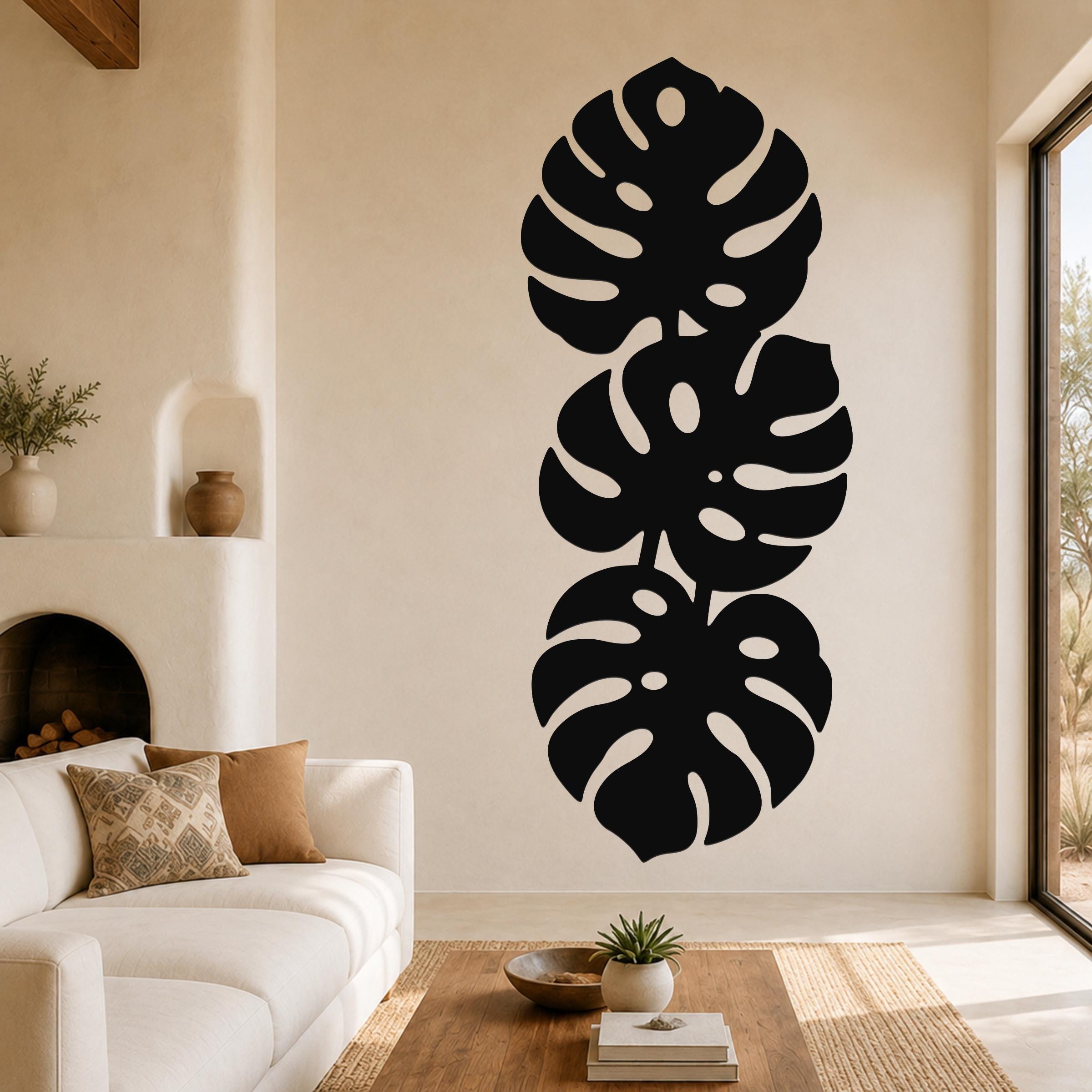 Black metal wall art featuring three stacked monstera leaf cutouts, mounted vertically on a living room wall above a white sofa.