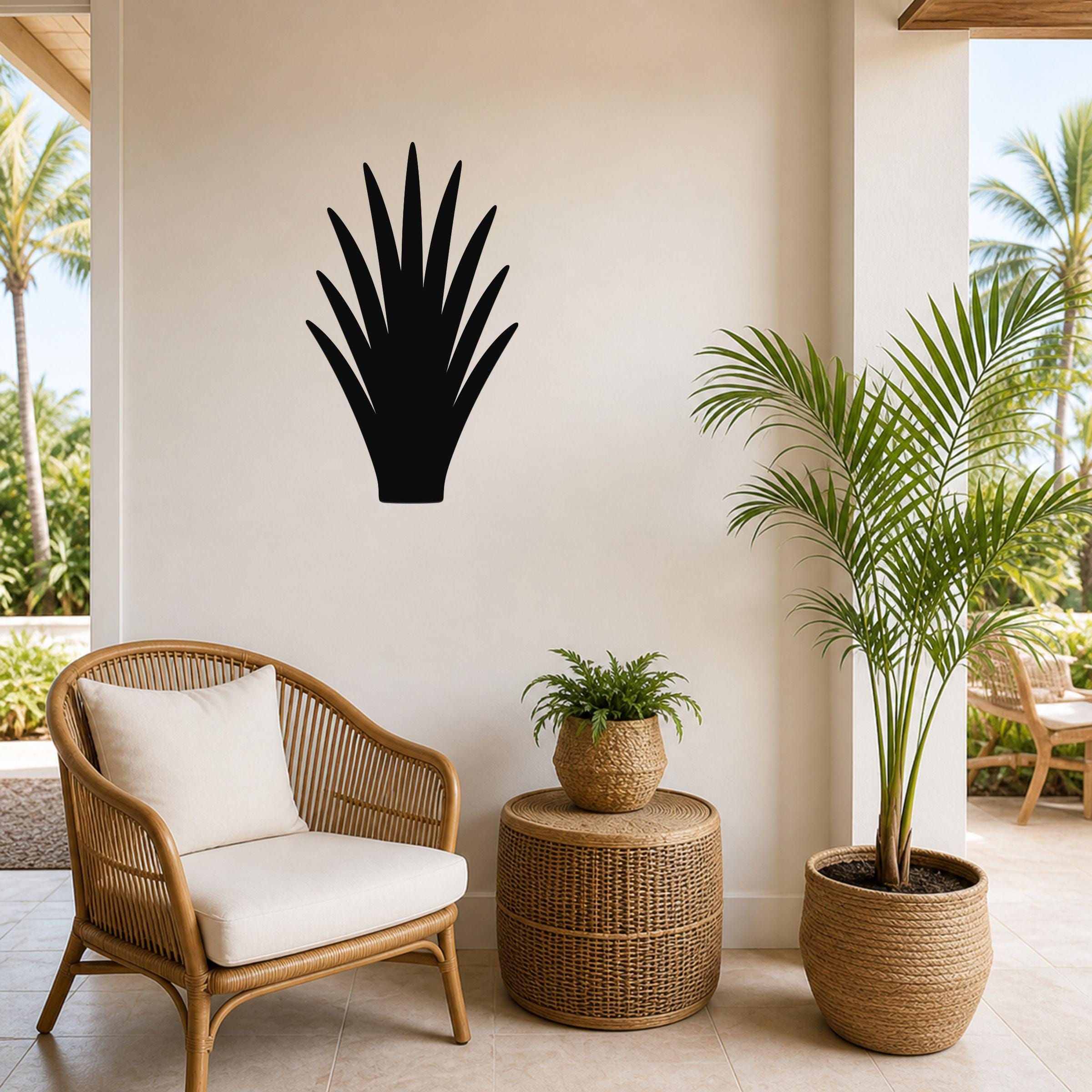 Black metal aloe vera wall art silhouette mounted on a light-colored wall above a wicker chair and potted plants in a patio setting.