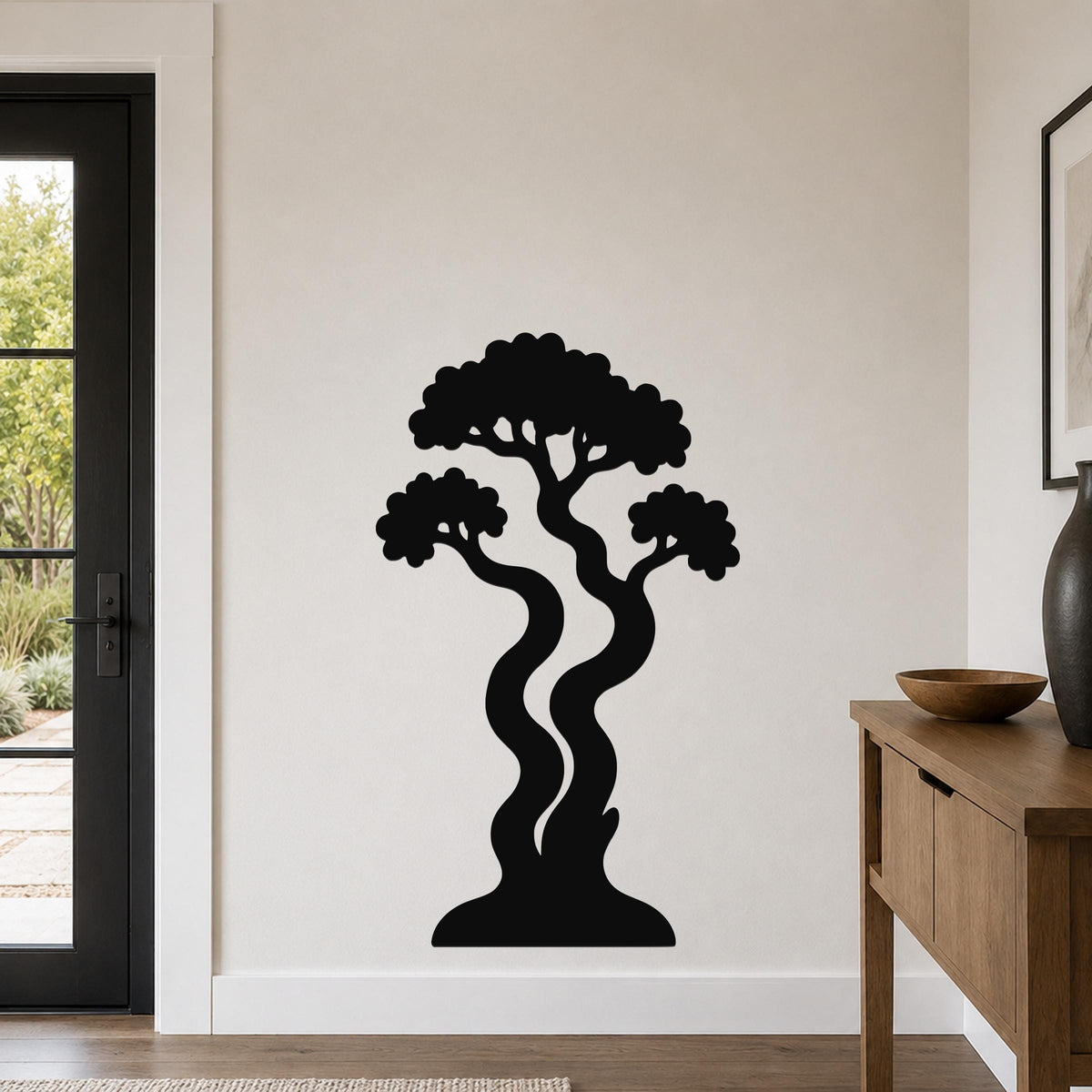 Black metal wall art in a bonsai tree silhouette with wavy trunks and layered canopy, mounted on a light-colored wall in a modern entryway.