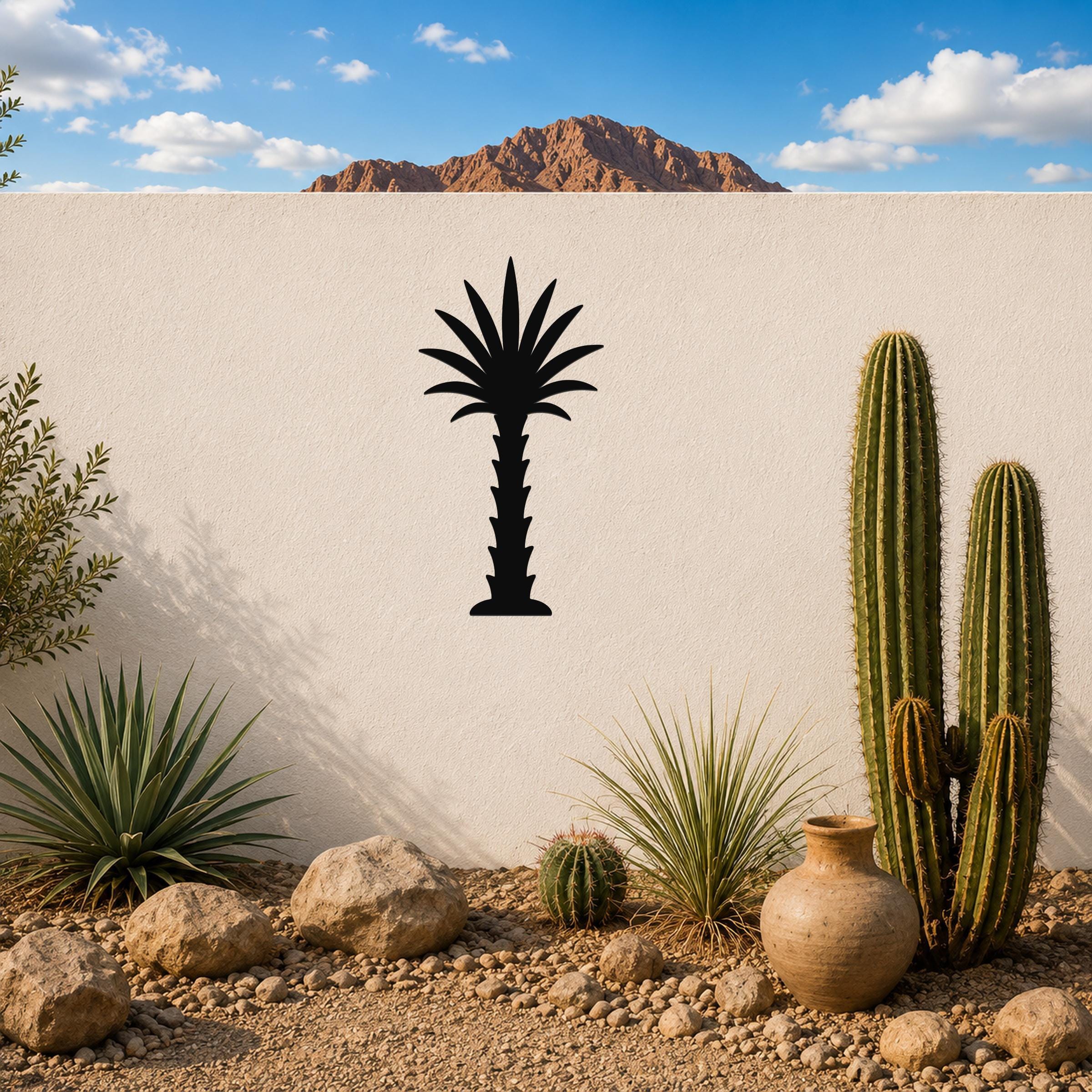 Black metal palm tree wall art silhouette mounted on a white outdoor wall above a desert garden with cacti and rocks.