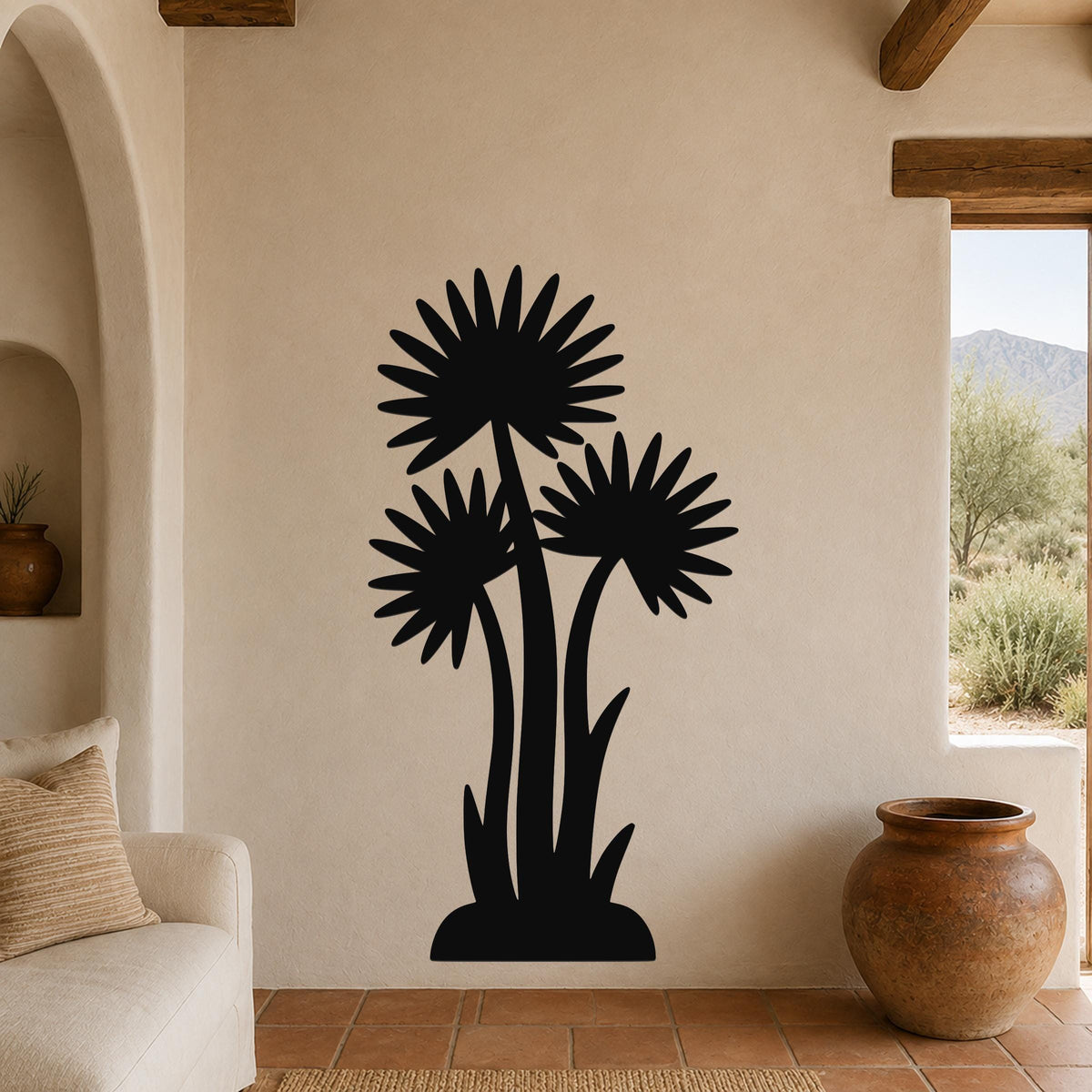 Black metal wall art featuring a silhouette of three desert yucca plants with spiky leaves, mounted on a beige stucco wall in a living room with terracotta flooring and wooden beams.