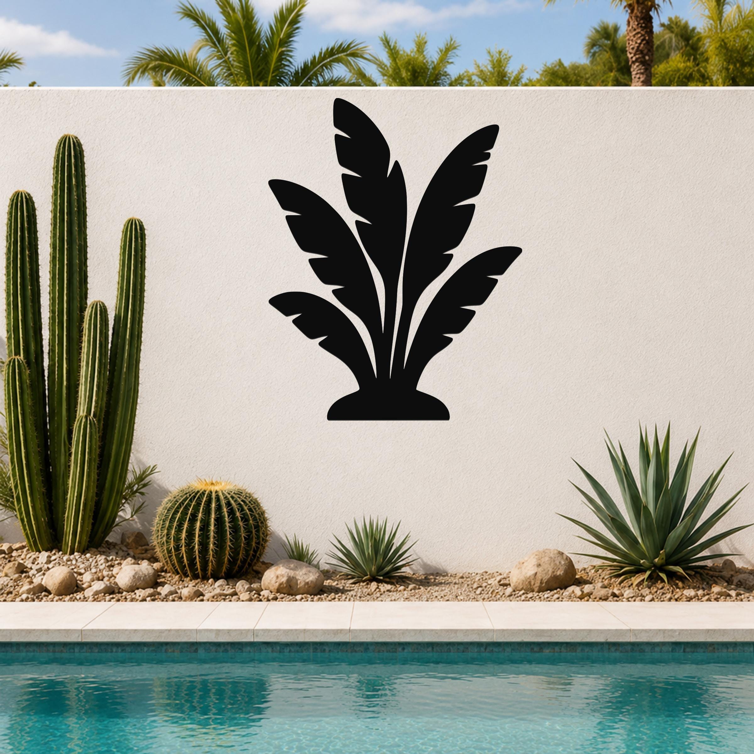 Black metal wall art in a tropical leaf silhouette design, mounted on a white outdoor wall beside a pool and desert plants.