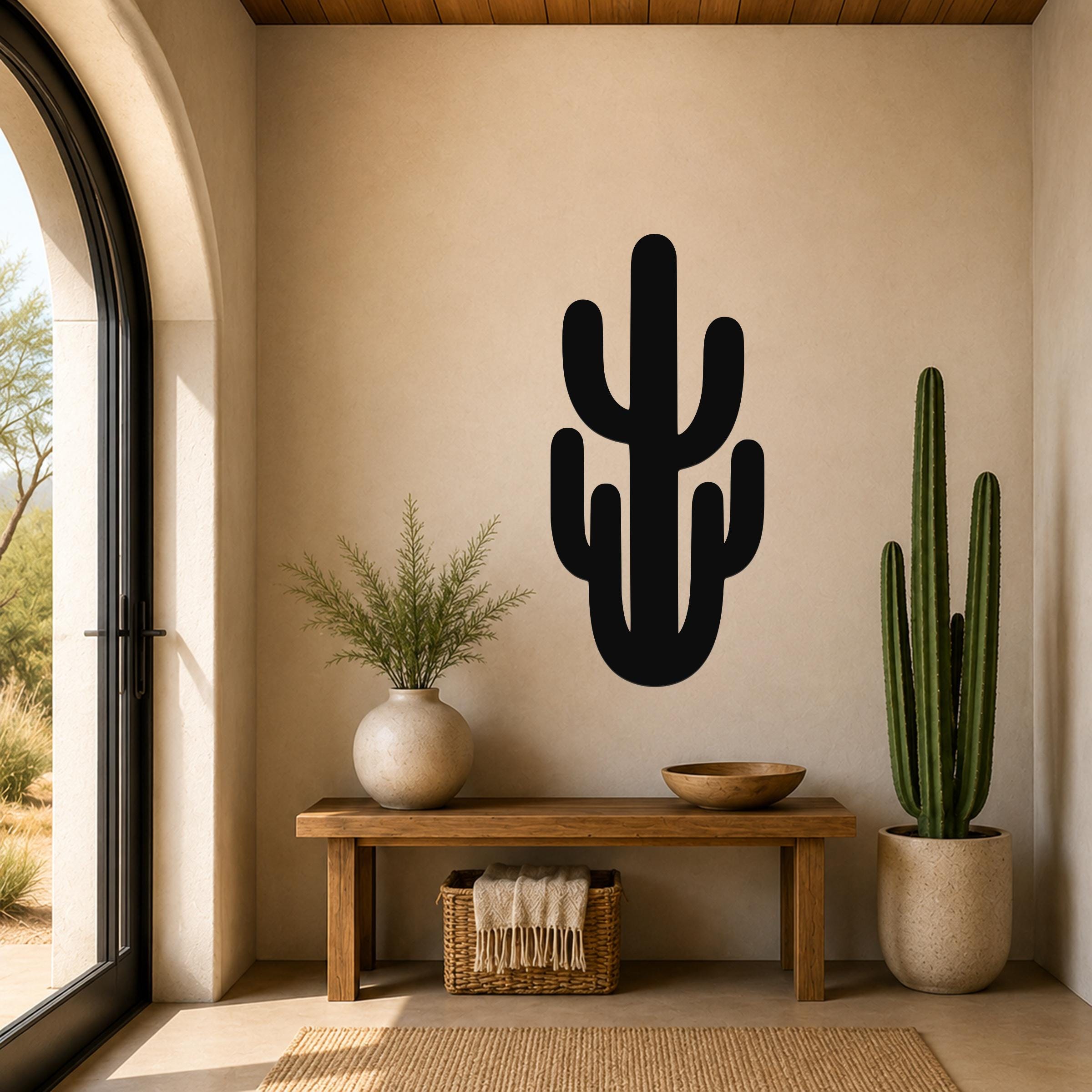 Black metal saguaro cactus wall art with cutout branches, mounted on a beige wall above a wooden bench in a sunlit entryway.