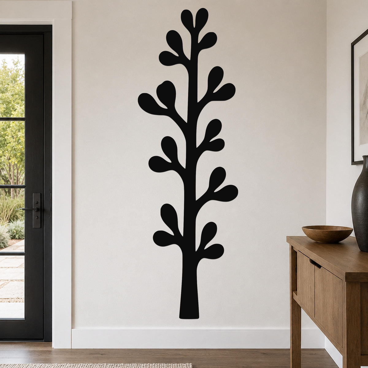 Black metal wall art in a tall, stylized olive branch silhouette with leaf cutouts, mounted on a living room wall above a wooden console table.