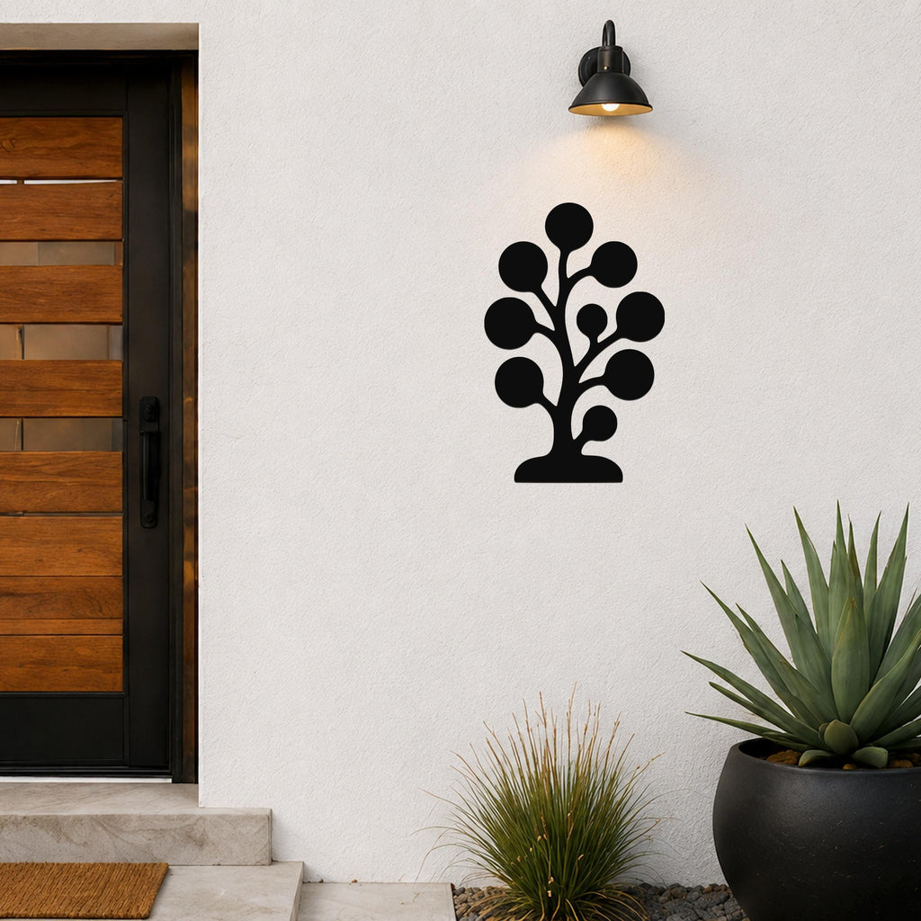 Black metal wall art in a tree silhouette with circular leaf cutouts, mounted on a white exterior wall beside a wooden door and potted plants.