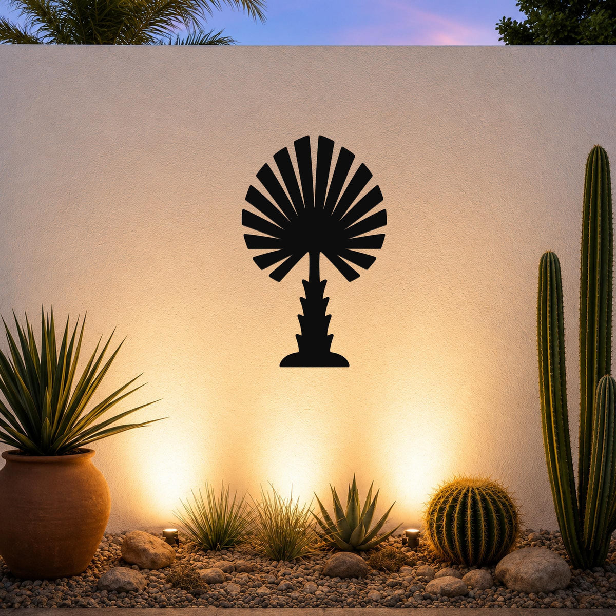 Black metal palm tree wall art with fan-shaped fronds, mounted on a textured outdoor wall above a desert garden with cacti and warm uplighting.