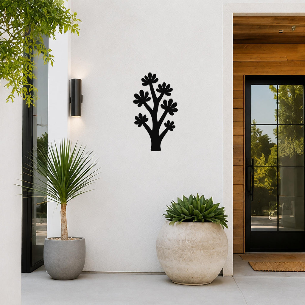 Black metal wall art in a tree silhouette with flower cutouts, mounted on a white outdoor wall beside a glass door and potted plants.