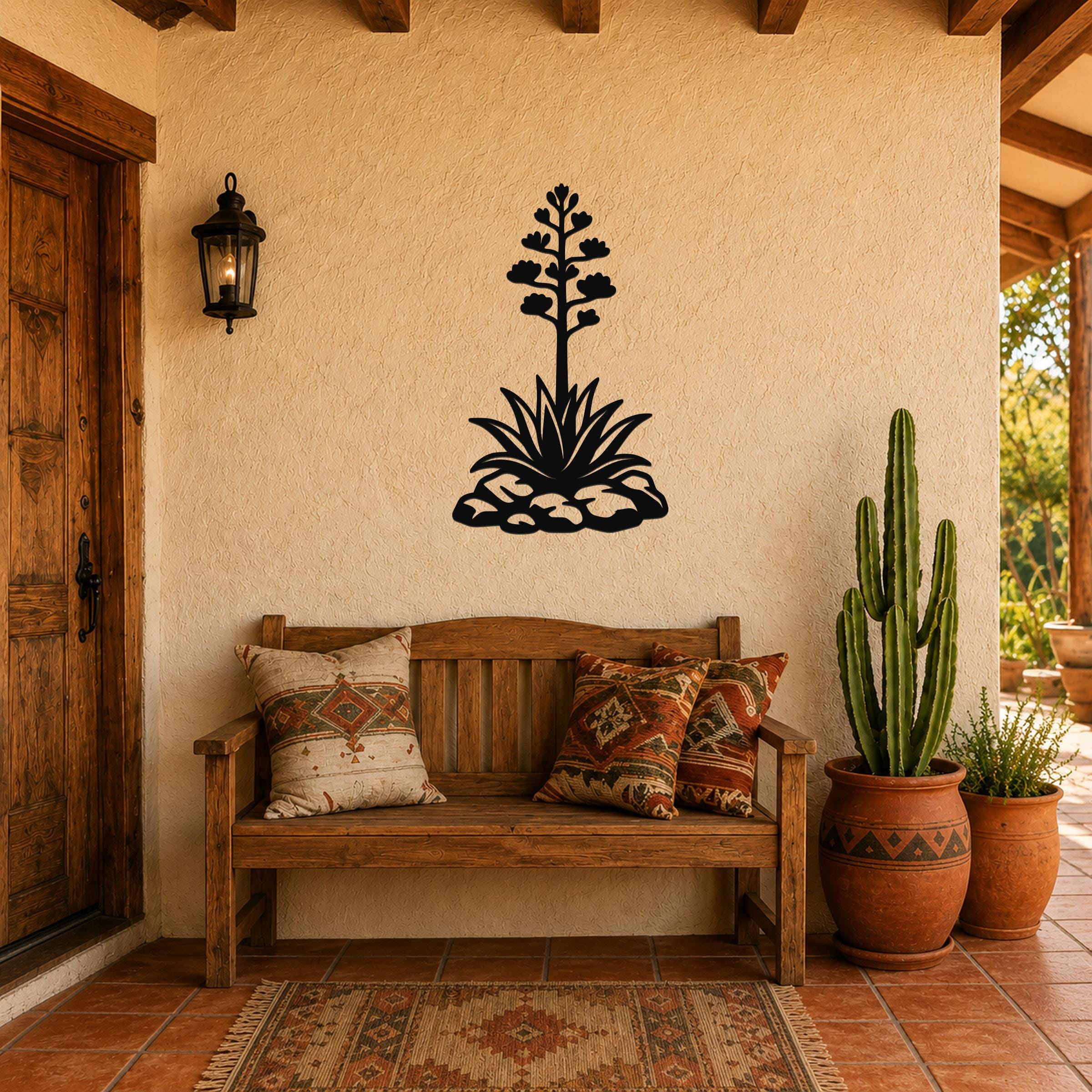 Black metal agave wall sculpture with blooming flower and leaf cutouts, mounted on a stucco wall above a wooden bench in a patio entryway.