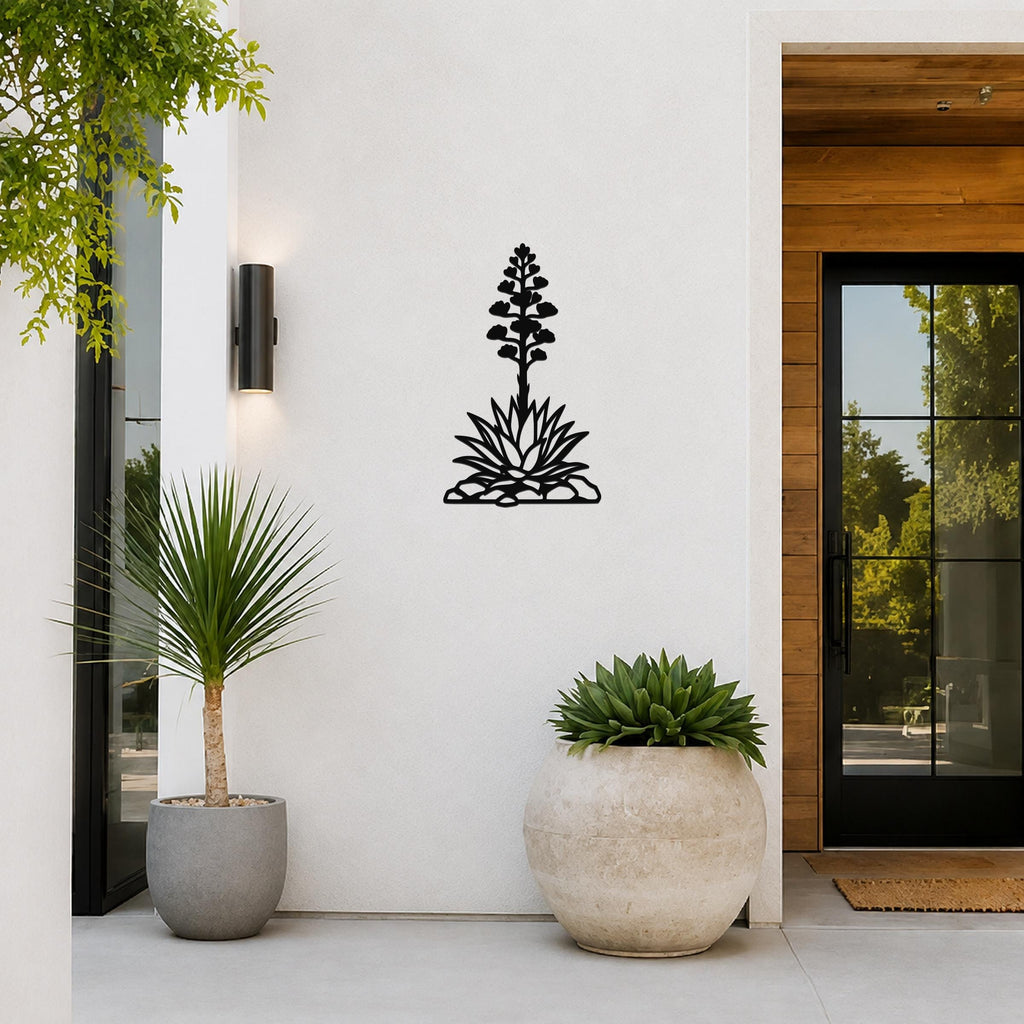 Black metal agave wall art with blooming flower and spiky leaves cutout, mounted on a white exterior wall beside a glass door and potted plants.