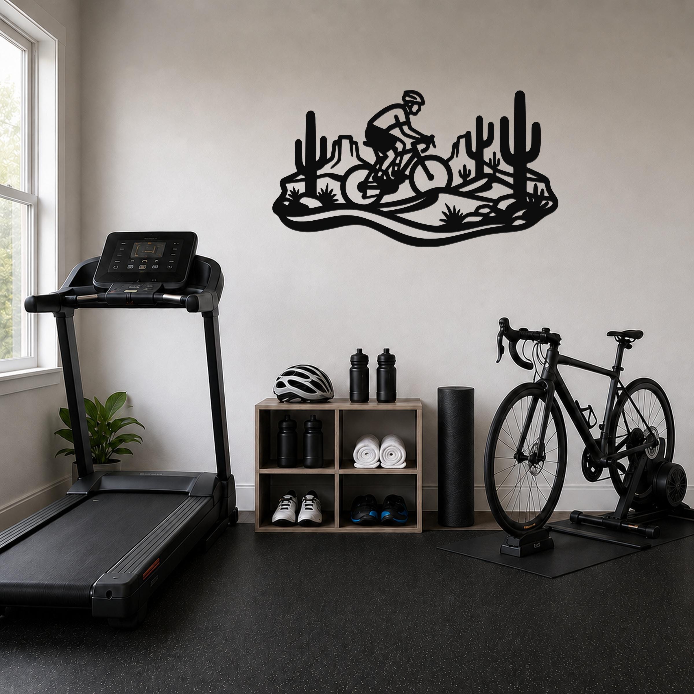 Black metal wall art featuring a cyclist silhouette on a mountain bike through a Sonoran desert scene with saguaro cacti, mounted on a wall above a storage shelf in a home gym.