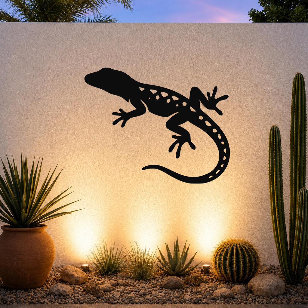 Black metal lizard wall art with internal cutout details, mounted on a textured outdoor wall above a desert garden with cacti and warm uplighting.