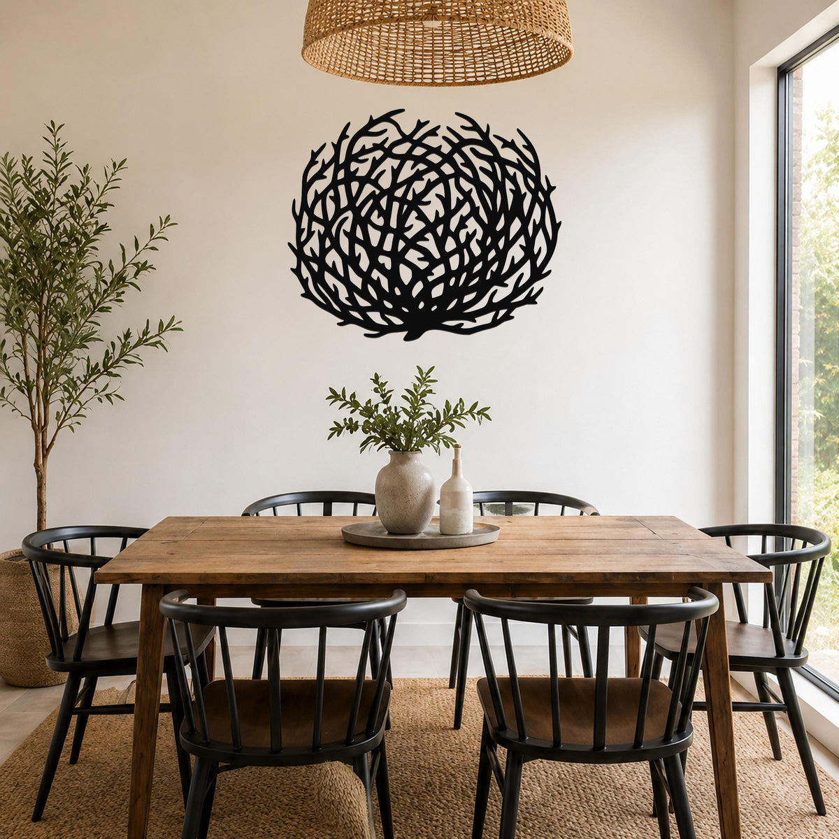 Black metal wall art in a circular tumbleweed silhouette with intricate branch cutouts, mounted on a white wall above a wooden dining table in a bright dining room.