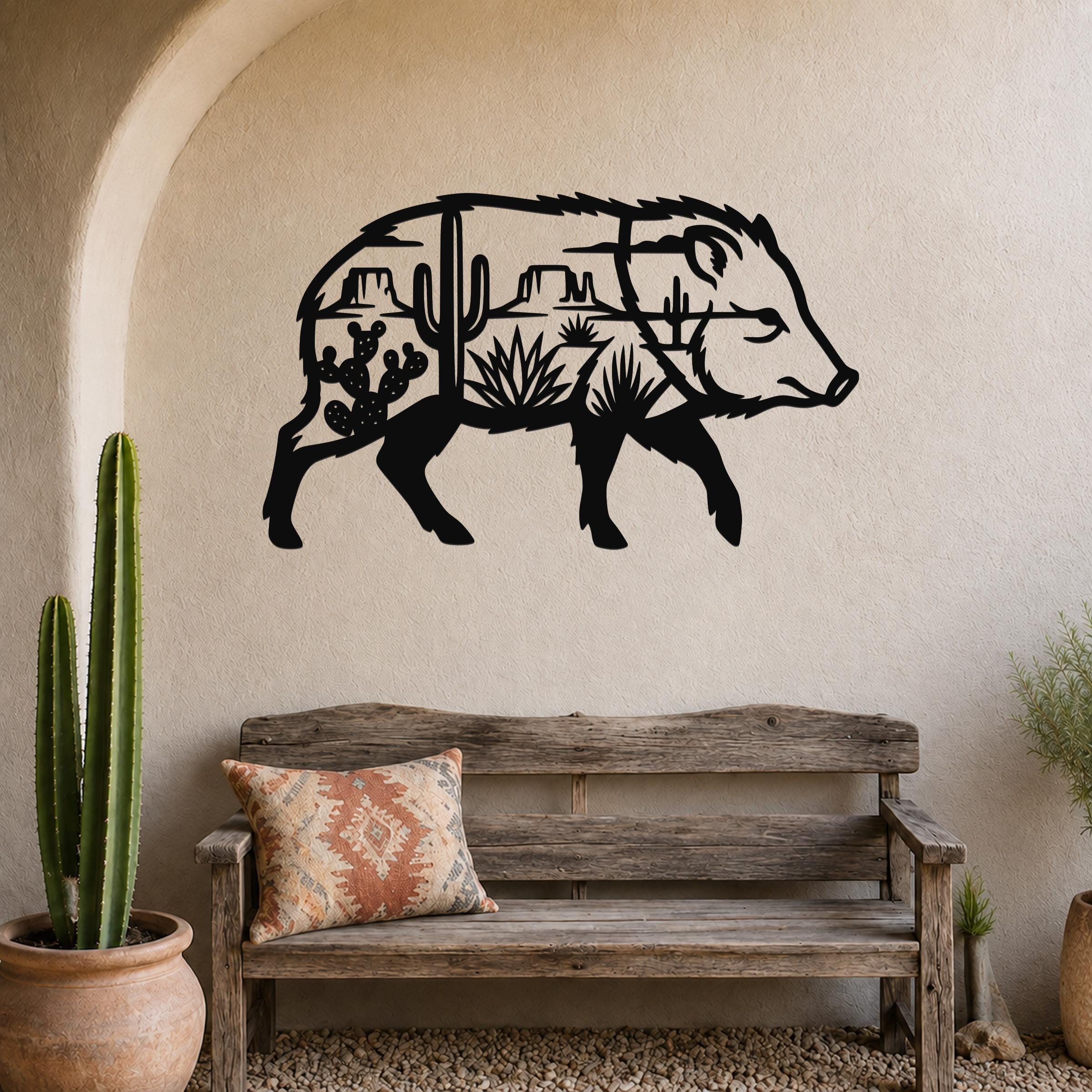 Black metal wall art in the silhouette of a javelina, with desert landscape cutouts including cacti and mountains, mounted on a beige wall above a wooden bench in a rustic living space.
