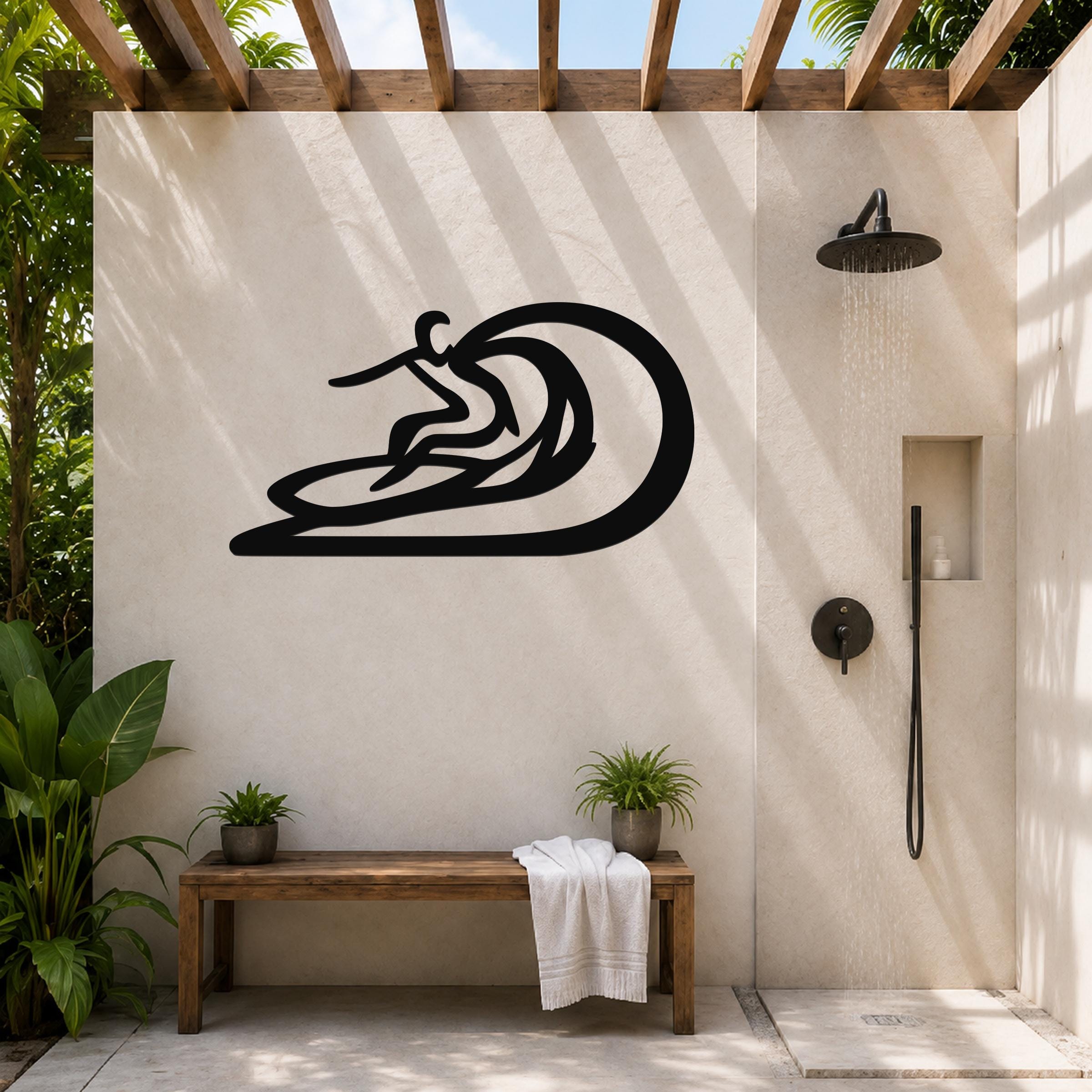 Black metal surfer wall sculpture with wave cutout, mounted on a beige outdoor shower wall above a wooden bench.