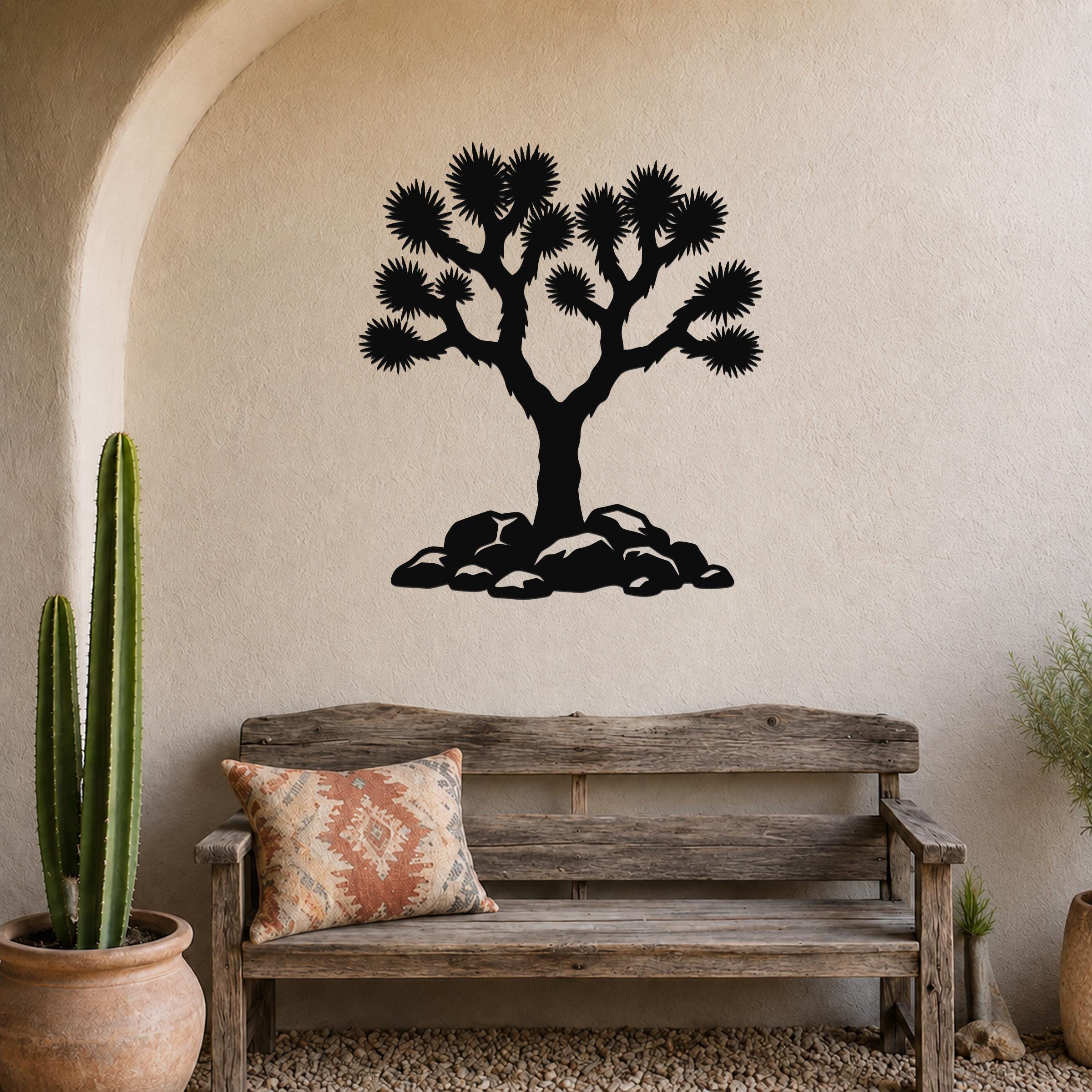 Black metal wall art silhouette of a Joshua tree with spiky branches and rocks at its base, mounted on a beige stucco wall above a wooden bench in a southwestern-style living space.