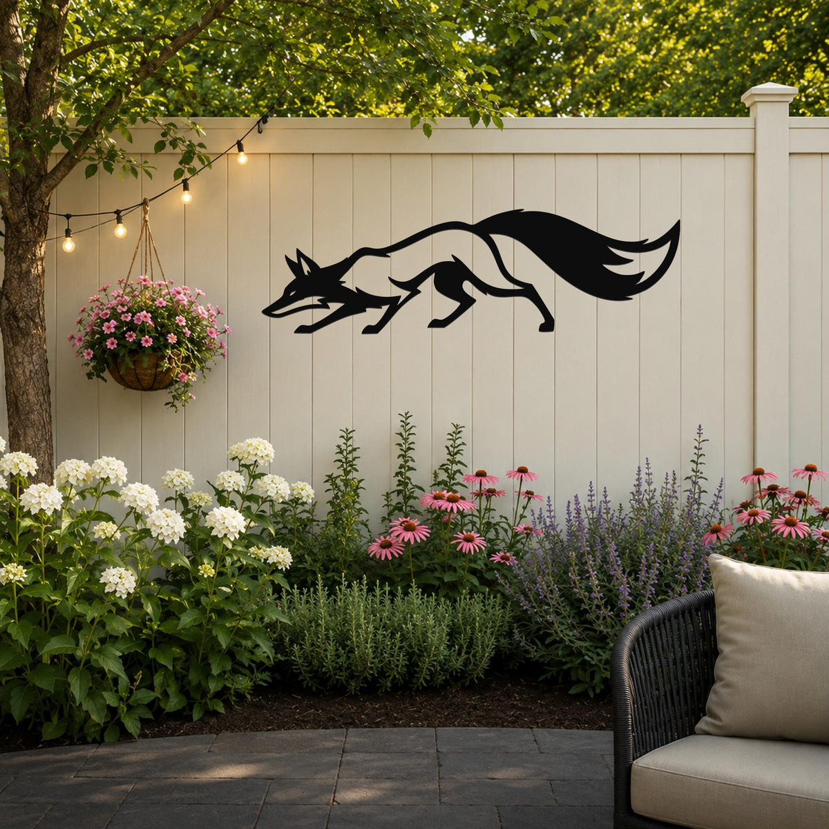 Black metal fox silhouette wall art mounted on a light beige outdoor fence, above a garden bed with white and pink flowers and a hanging basket of pink blooms.