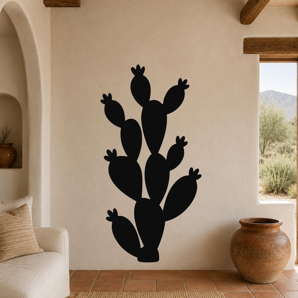 Black metal prickly pear cactus wall art with multiple segmented pads and small flower cutouts, mounted on a beige stucco wall in a living room with terracotta flooring and wooden beams.