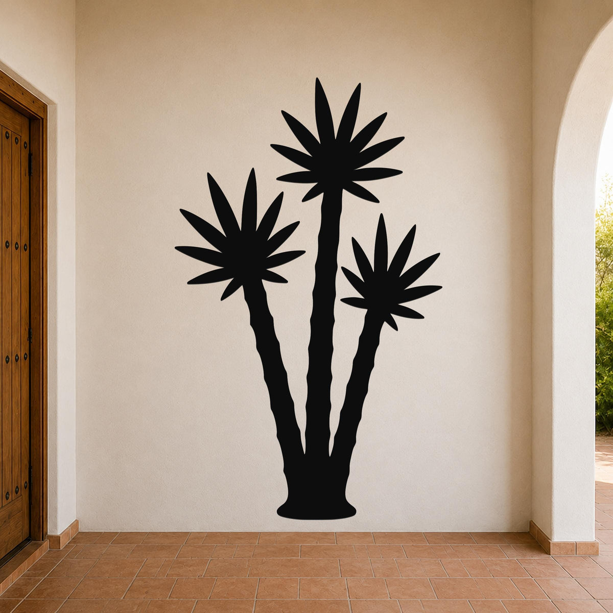 Black metal wall art featuring a silhouette of three abstract desert plants, mounted on a beige wall in a hallway with terracotta flooring.