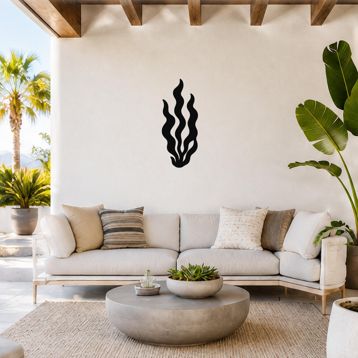 Black metal kelp wall art with flowing seaweed cutouts, mounted on a white wall above a light-colored sofa in a bright outdoor living space.