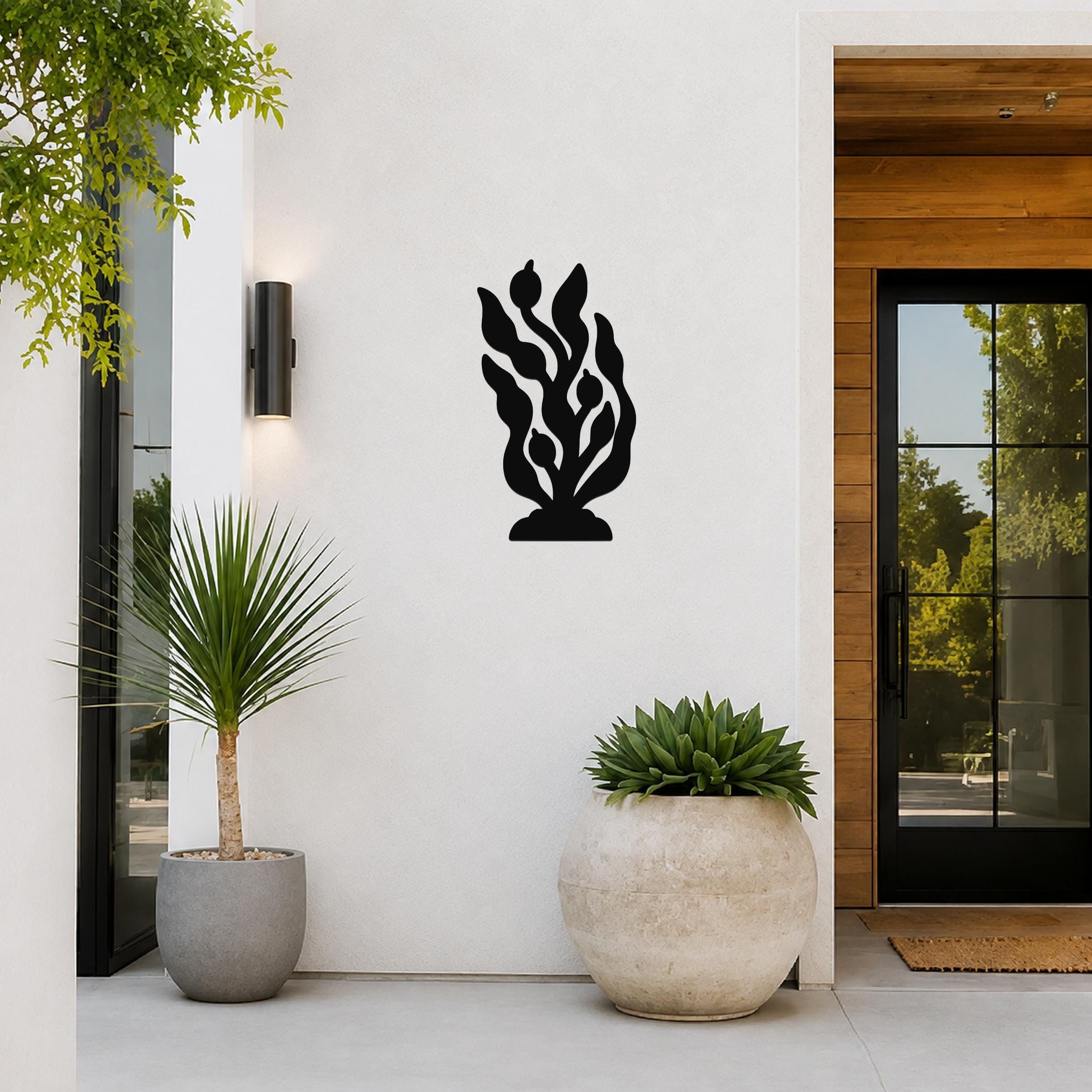 Black metal kelp and seaweed wall art with leafy cutouts, mounted on a white exterior wall beside a glass door and potted plants.