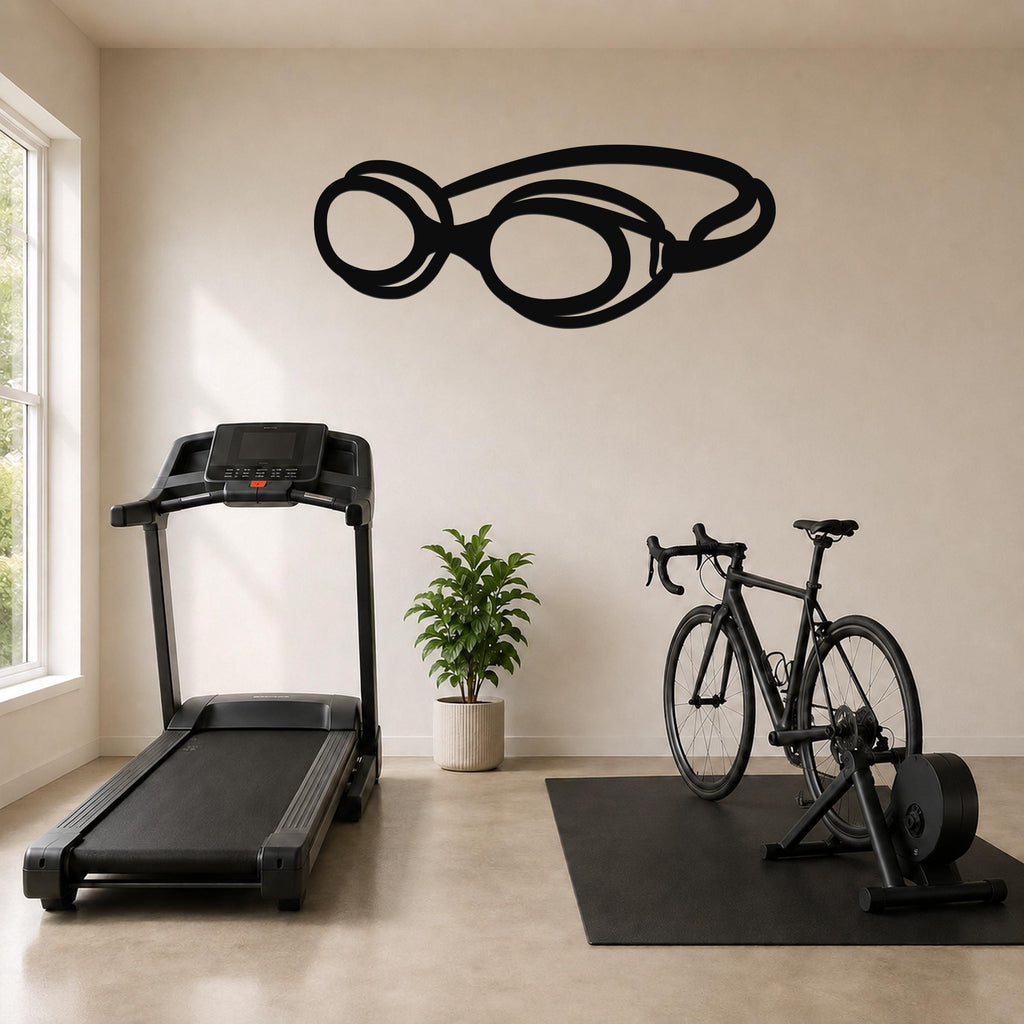 Black metal swim goggles wall art silhouette mounted on a beige wall above a treadmill and exercise bike in a home gym.