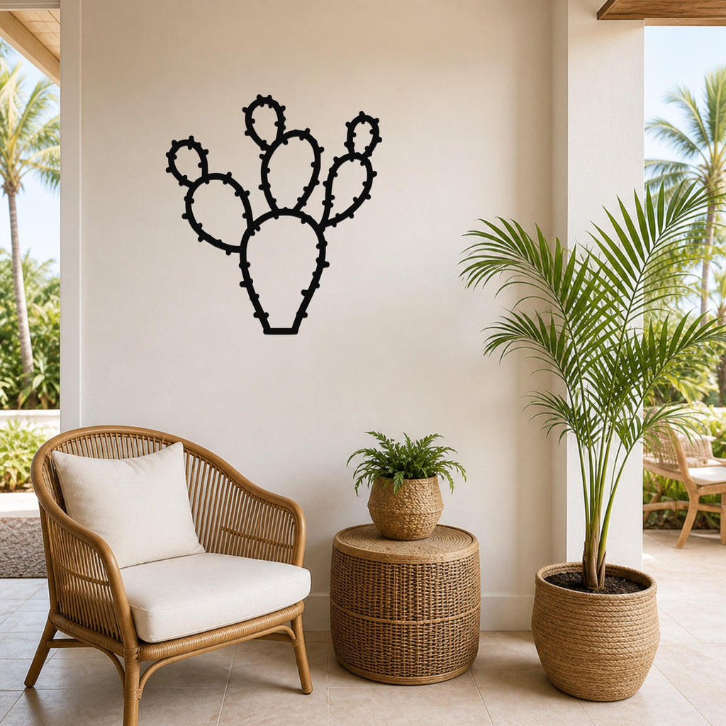 Black metal wall art in the silhouette of a prickly pear cactus with detailed spine cutouts, mounted on a light-colored wall above a wicker chair in a patio setting.