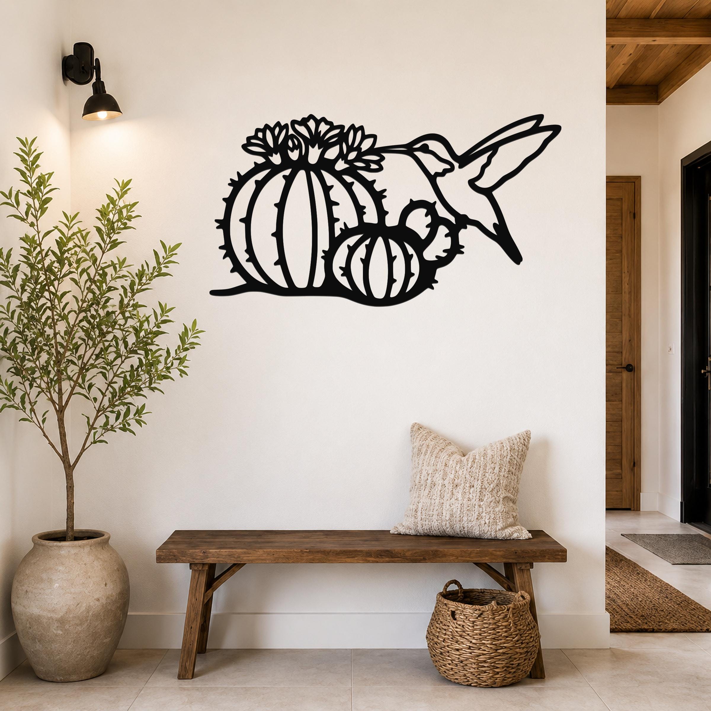 Black metal wall art featuring a hummingbird and barrel cactus with flower cutouts, mounted on a white wall above a wooden bench in a hallway.