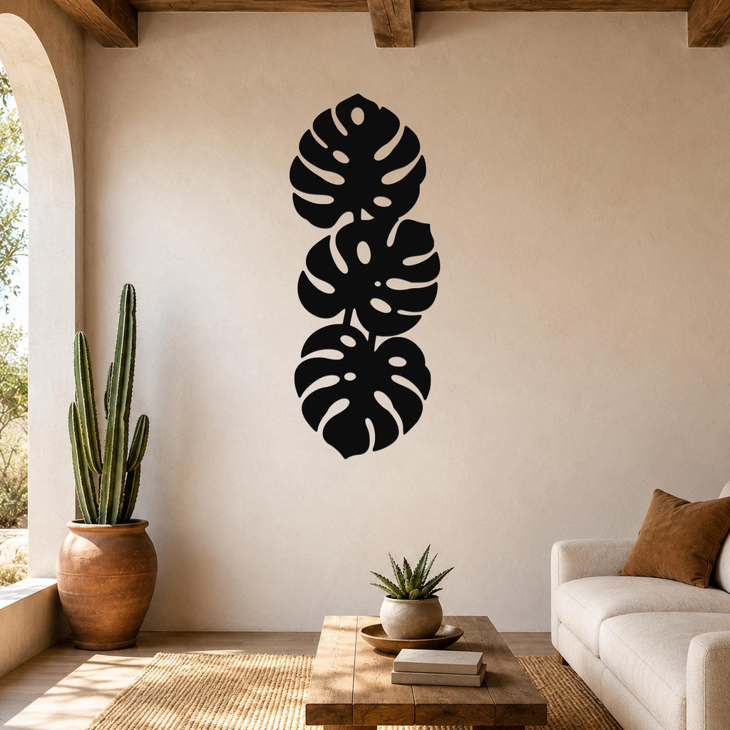 Black metal wall art featuring three stacked monstera leaf cutouts, mounted vertically on a living room wall above a wooden coffee table.