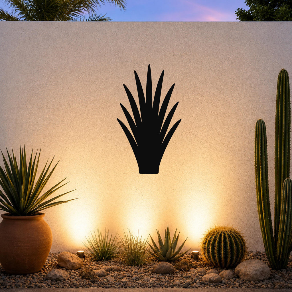 Black metal agave wall art silhouette mounted on a white outdoor wall above a desert garden with cacti and warm uplighting.