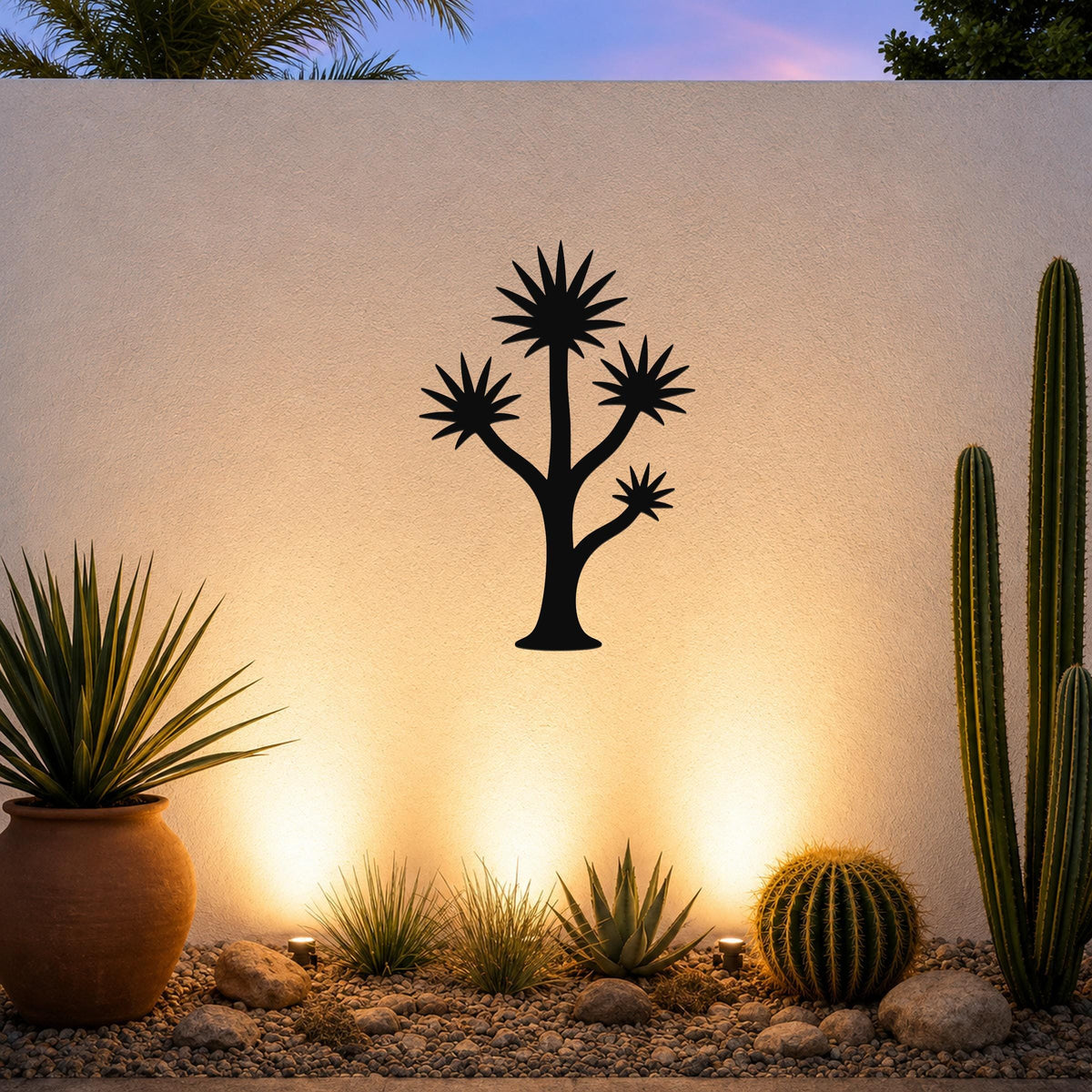 Black metal wall art in a yucca tree silhouette with starburst leaves, mounted on a white outdoor wall above a desert garden with cacti and warm uplighting.