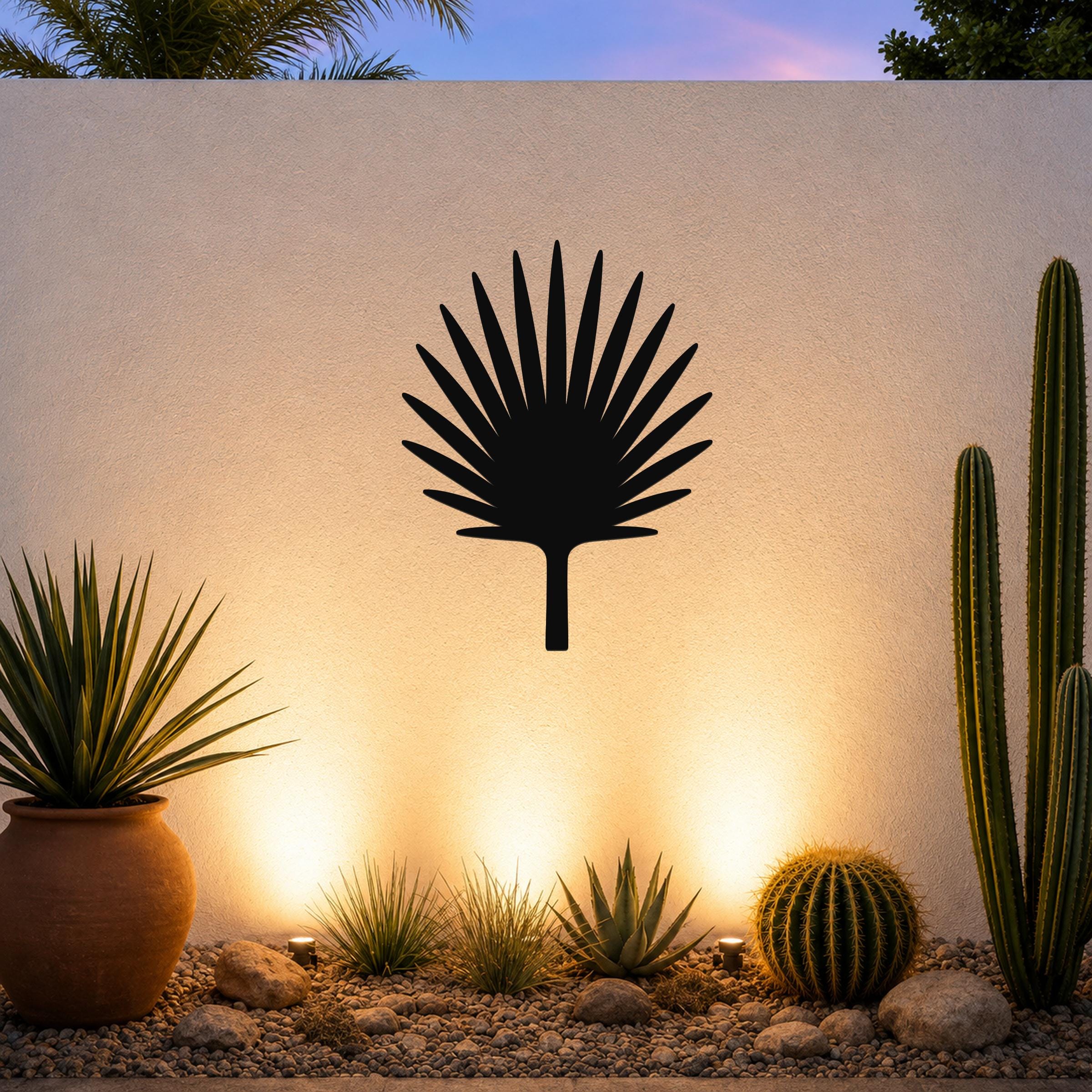 Black metal fan palm tree wall art silhouette mounted on a textured outdoor wall above a desert garden with cacti and warm uplighting.
