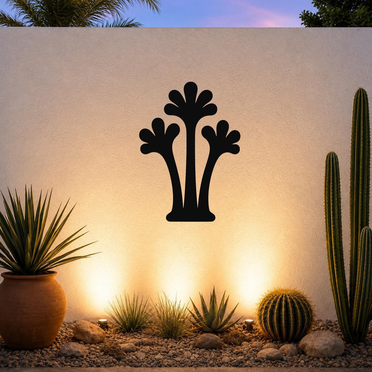 Black metal palm tree wall art with three frond silhouettes, mounted on a white outdoor wall above a desert garden with cacti and warm uplighting.